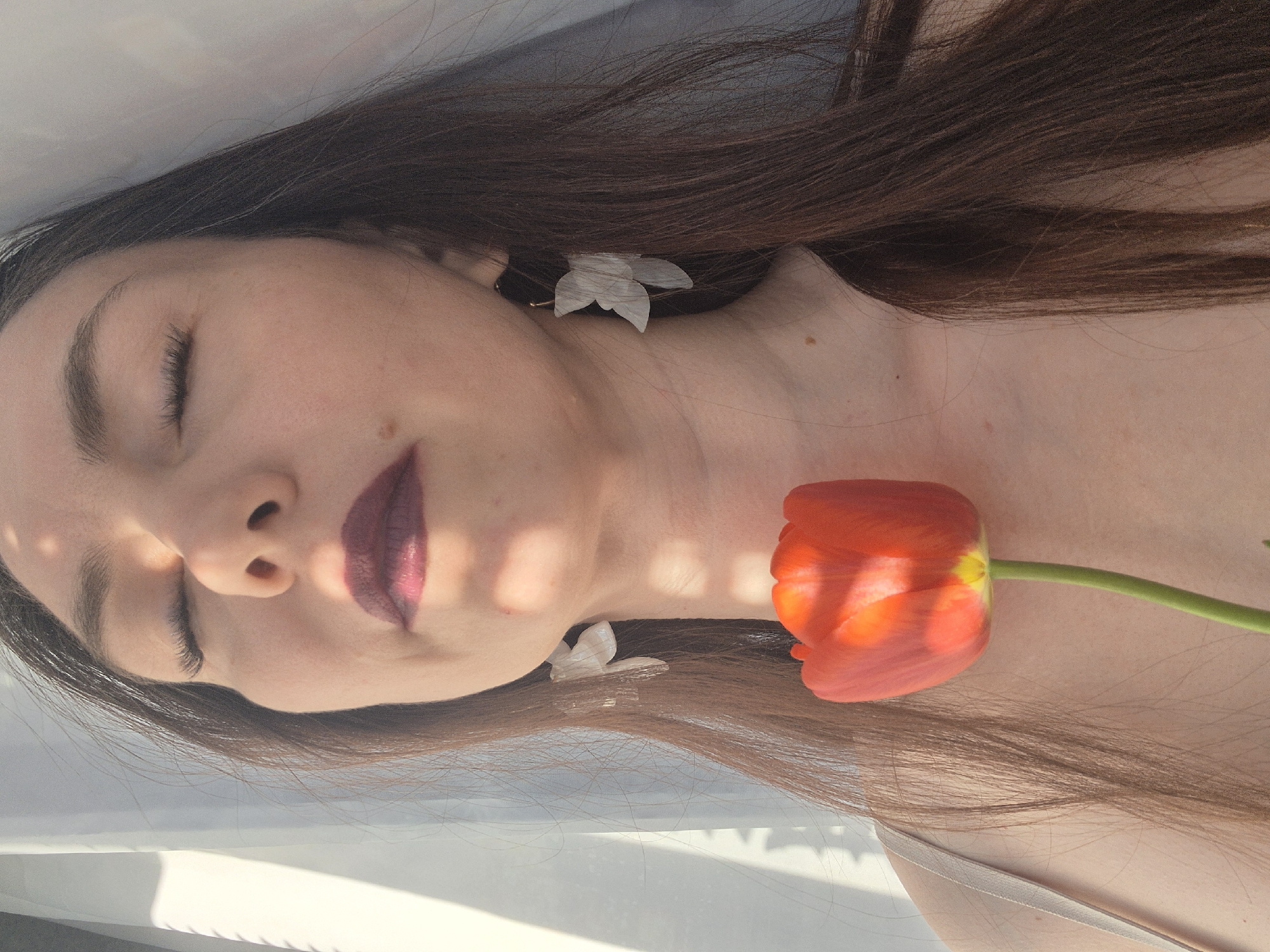 A young woman lies on her back in an indeterminate indoor location, likely a private residence. She has dark brown hair, fair skin, and is wearing a dark lipstick. Her eyes are closed, and she appears to be resting. A single red tulip lies against her neck. She is wearing white, flower-shaped earrings. There is light, possibly sunlight, filtering through a nearby window, casting shadows across her face. The setting is ambiguous, but it is likely a staged or artistic portrait.