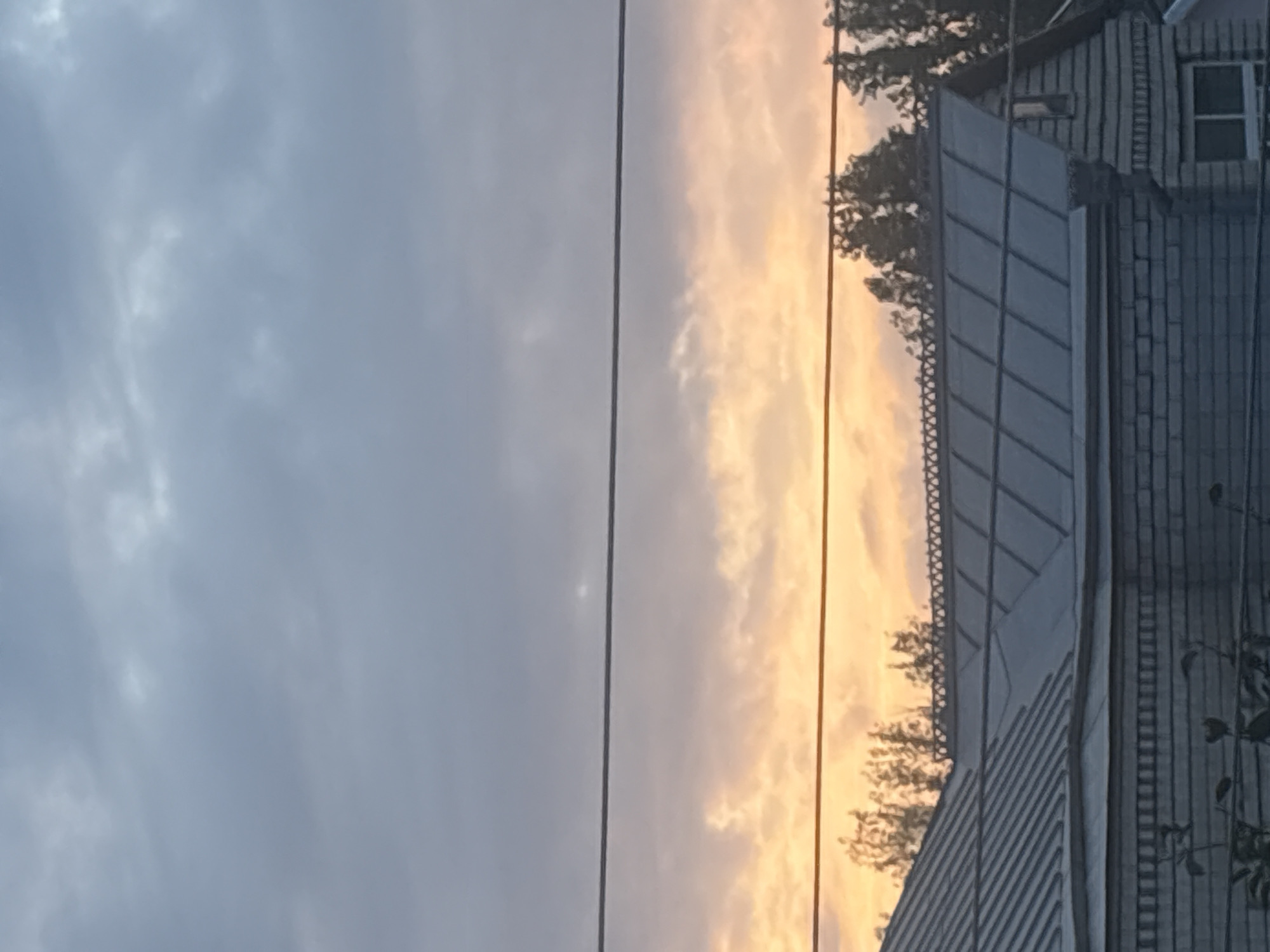 The image depicts a suburban scene at dusk, focusing on the roofs of buildings against a cloudy sky. The sky transitions from a pale blue-grey at the top to hues of orange and yellow closer to the horizon, suggesting a sunset. Two horizontal power lines cross the frame, dividing the sky. Below the sky, the upper parts of two connected structures are visible. The structure in the foreground has a multi-pitched, dark-colored metal roof with visible horizontal seams. It appears to be a house with brick siding. The roof of the adjacent structure, slightly further back and to the right, is also visible, featuring a similar metal roofing material and a peaked section with a decorative trim along the roofline. Silhouetted trees are visible between the sky and the buildings, with some branches extending above the decorative roof trim. The overall impression is one of a quiet, residential area at the end of the day. The specific location in Georgiyevsk, Russia, is not explicitly discernible from the visual elements of the image alone.