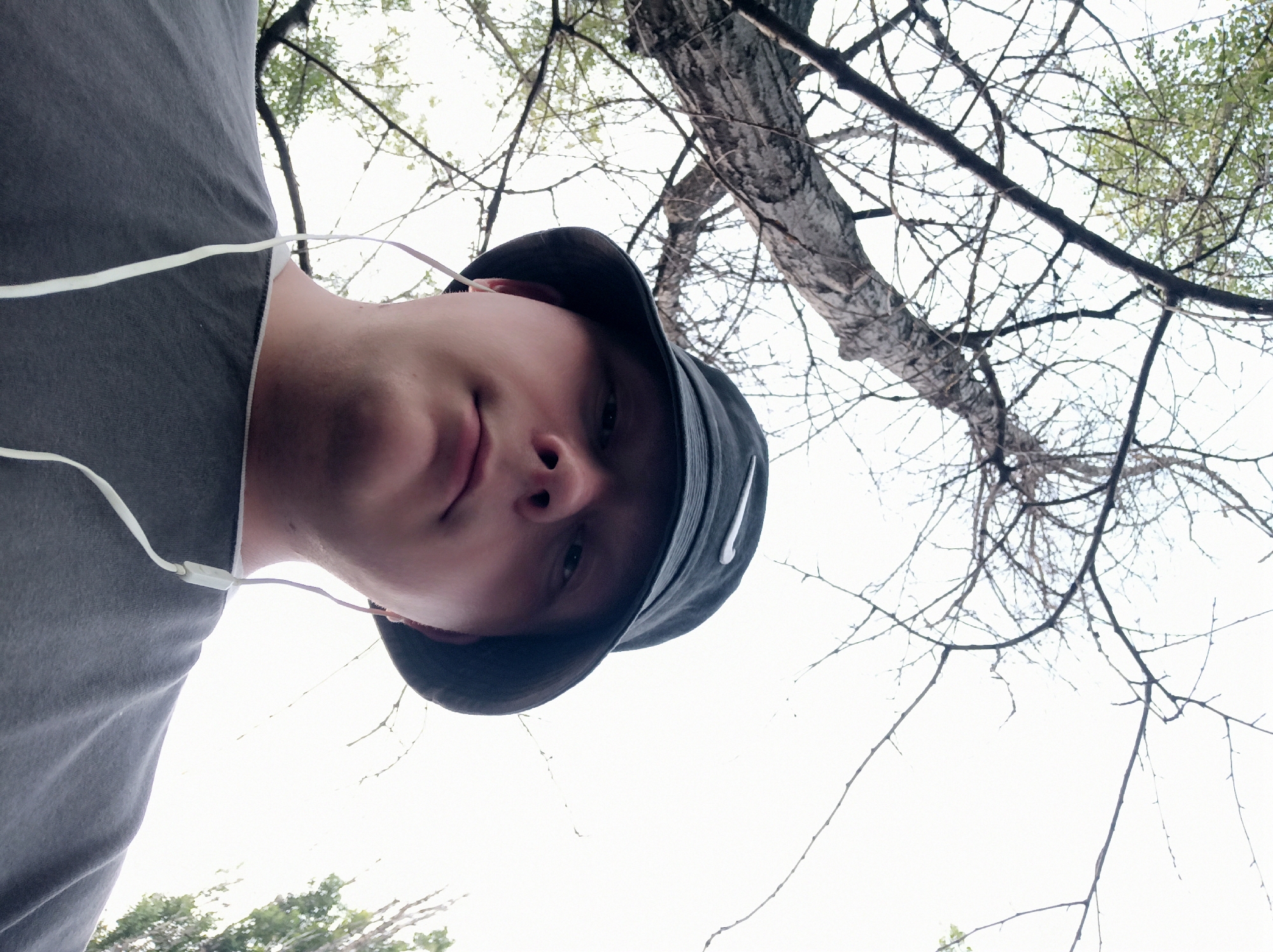 A person wearing a dark bucket hat with a white logo and white earbuds is shown from a low angle. The person is looking upwards. The background consists of a large tree with bare branches and some leaves against a bright, overcast sky. The person is wearing a gray t-shirt.