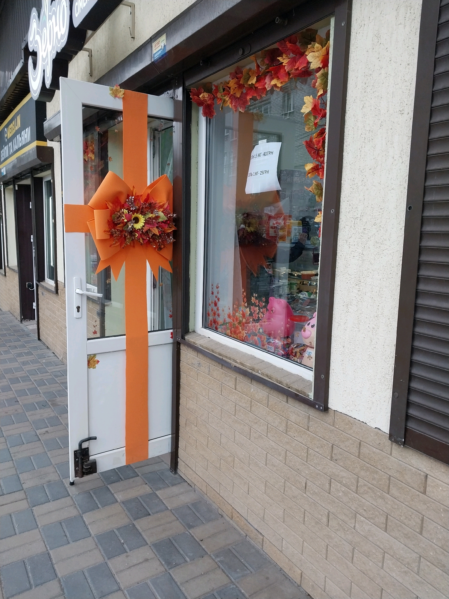The image shows the exterior of a shop in Dnipro, Ukraine. The shop has a large glass window decorated with artificial autumn leaves. A white door is partially open, decorated with a large orange bow and artificial autumn flowers. The shop is located on a sidewalk paved with interlocking gray and beige concrete pavers. The building facade is covered with a textured cream-colored plaster and has dark brown trim around the window. A dark brown security shutter is visible on the right side of the image. The shop's signage is visible above the door, with the word "ЗЕРНО" (which translates to "GRAIN") in large, stylized white lettering. Below this, there is smaller black lettering that is partially obscured. There are no people visible in the image.