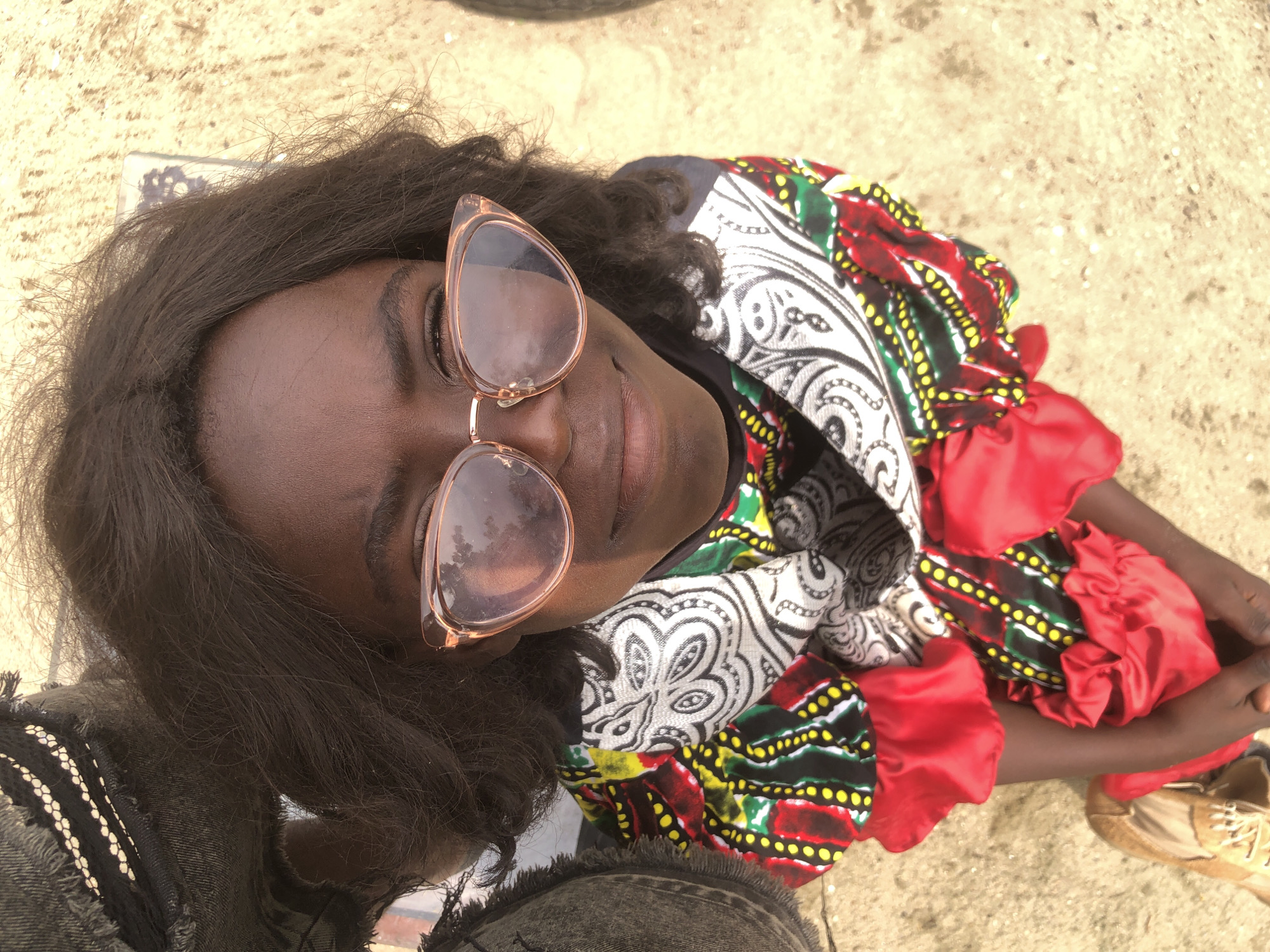 A high-angle, close-up, full-body shot shows a young Black girl looking up at the camera, smiling slightly. She is wearing cat-eye sunglasses with rose-gold frames, and her dark, curly hair frames her face. Her outfit consists of a patterned top with a black, white, and red African print fabric, featuring yellow polka dots and red ruffled sleeves. She is also wearing what appears to be a black shirt underneath the patterned top. To her left, a dark denim garment with frayed edges is visible, possibly part of jeans or a skirt. To her right, part of a shoe with laces is visible. The ground beneath her is a sandy, light-brown surface, typical of outdoor areas in Ado, Nigeria. The lighting suggests it is daytime.