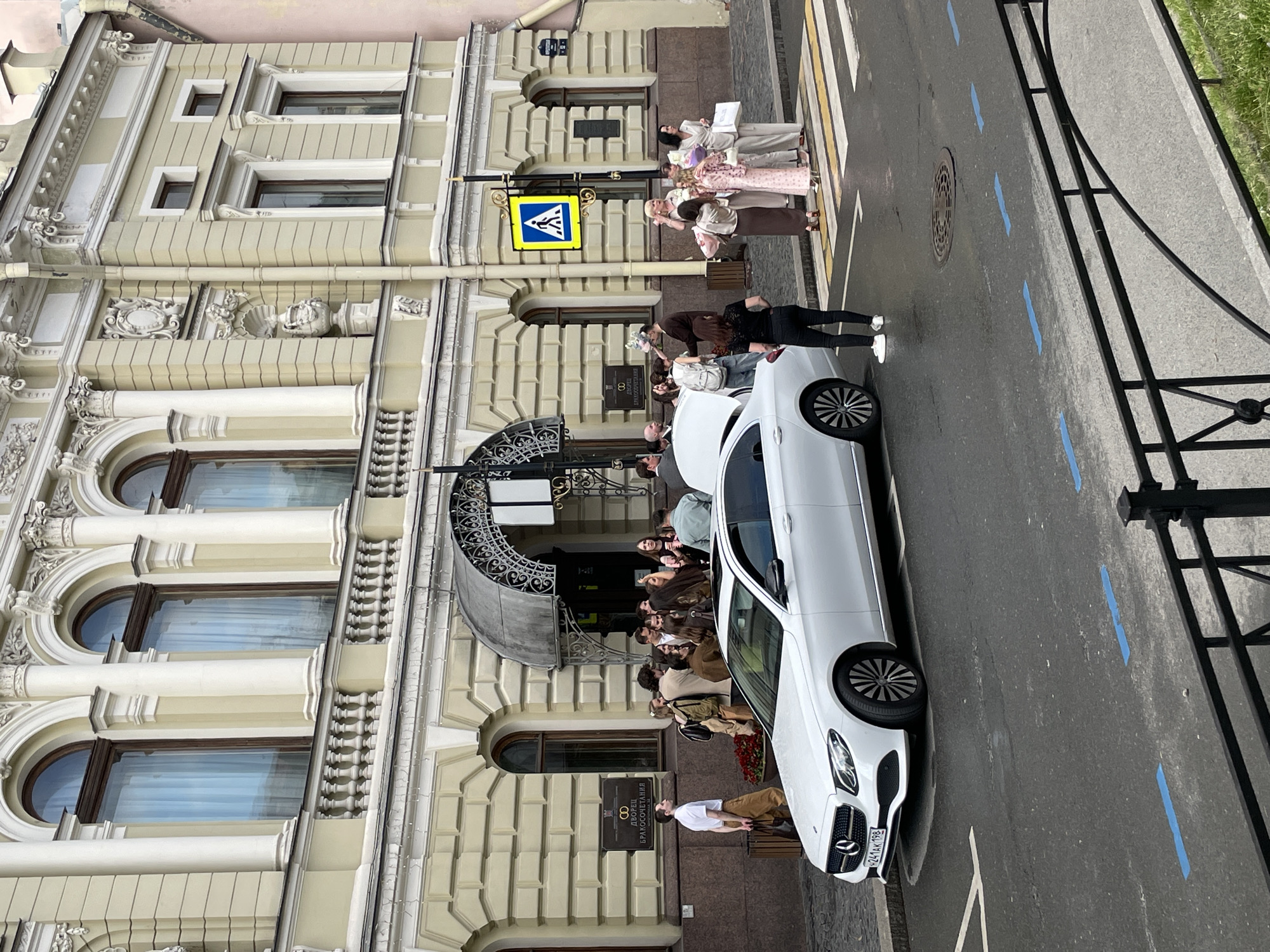 The image depicts a vibrant daytime street scene in Saint Petersburg, Russia, in front of a grand, ornate building. The multi-story edifice is characterized by its light-colored facade, classical architectural elements including pilasters, cornices, arched windows with decorative keystones, and elaborate relief carvings.
In the foreground, a white Mercedes-Benz sedan is parked alongside the curb. A group of people, dressed in smart casual to semi-formal attire, are gathered around the car and near the building's entrance, which features an elegant wrought iron canopy. Another group of people is visible further down the sidewalk, near a pedestrian crossing. The general activity suggests a special occasion, likely a wedding, given the visible sign.
The street features a zebra crossing and several blue rectangular markings painted on the asphalt, along with a manhole cover. A yellow pedestrian crossing sign, displaying a black human figure, is mounted on a pole near the crosswalk. To the right, a black metal fence separates the street from a patch of green grass.
A prominent sign on the building entrance clearly displays "ДВОРЕЦ БРАКОСОЧЕТАНИЯ" (Palace of Marriage/Wedding Palace) above a symbol of intertwined rings, confirming the venue's purpose. Below this, in smaller text, "ОСОБНЯК ПОЛОВЦОВА" (Polovtsov Mansion) identifies the historical name of the building. The license plate of the white car, "У241КХ 198", is also visible, with "198" being the region code for Saint Petersburg. The scene suggests a pleasant day with people celebrating or preparing for an event.
