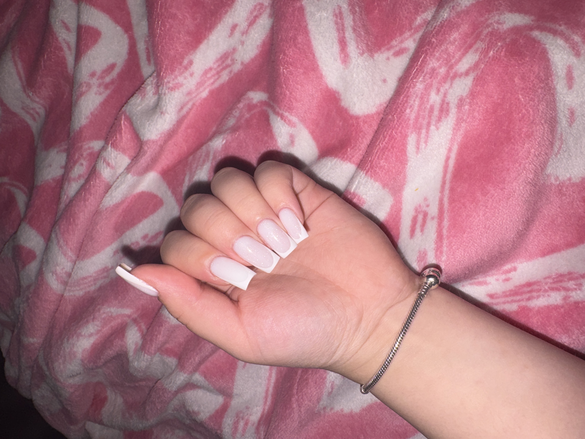 The image displays a person's left hand and lower forearm resting on a soft, patterned blanket. The hand has a light skin tone. The nails are long, with a square or coffin shape, featuring a milky white or very light pink base with prominent white tips, suggesting a modern French manicure style, possibly with a dip or ombre effect. Some of the nails also exhibit a subtle sparkle or shimmer. A silver-colored snake chain bracelet with a rounded charm or clasp is worn on the wrist.
The background is a plush blanket, predominantly pink with broad, abstract white patterns, possibly resembling brushstrokes or wavy lines. The fabric appears soft and slightly wrinkled or folded. The lighting in the image suggests an indoor setting, likely under artificial light, as indicated by the shadows and highlights on the hand and the bracelet. The image solely focuses on the hand and its immediate surroundings, presenting the nails and bracelet. No specific activity beyond posing the hand is discernible. No text is visible in the image. This scene is contextualized as being in Saint Petersburg, Russia.