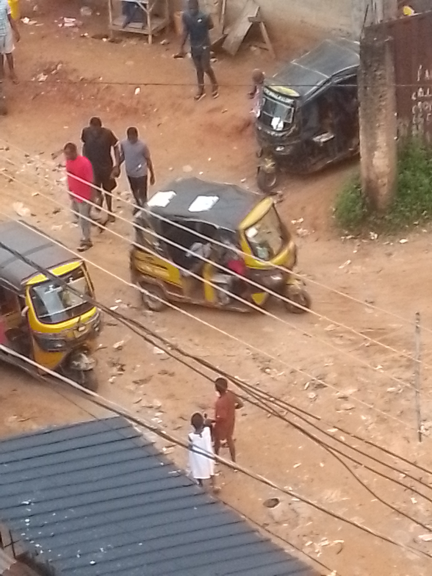 In Nkpor, Nigeria, a sandy, uneven road is lined with power lines and debris. Two auto-rickshaws, one yellow and black, the other dark, are present. Several people are on the road, including a person in a red shirt, a person in a black shirt, and a person in a gray shirt. Two children are also visible; one is wearing a white dress, and the other is in an orange outfit. In the background, there are structures, including a metal frame and a building. The overall scene suggests a busy street environment with active transportation and pedestrian movement.