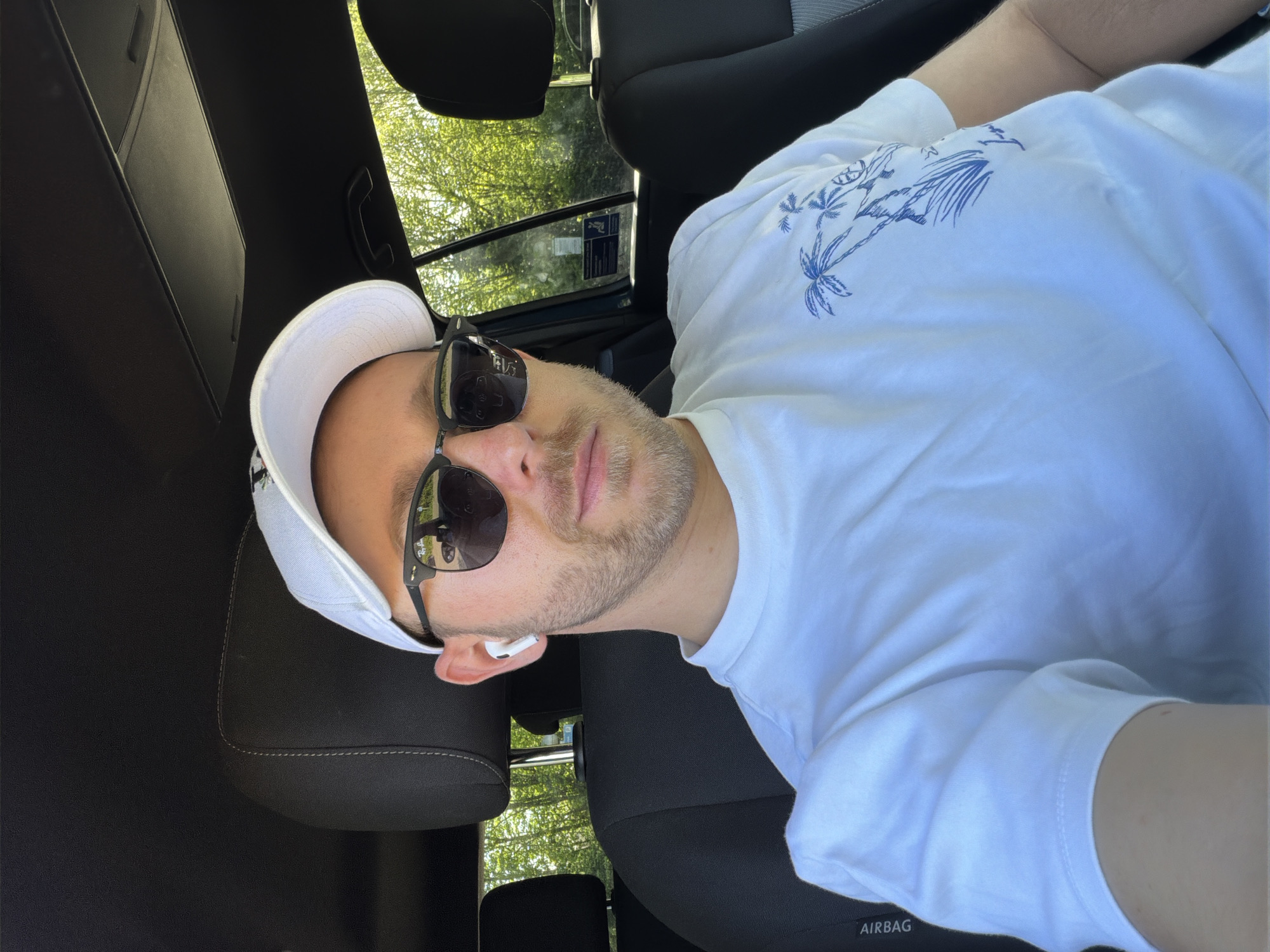 A selfie of a man in a car. He is wearing a white baseball cap, sunglasses, and a white T-shirt with a blue palm tree design. He is wearing an in-ear wireless headphone. The car is parked, and trees are visible through the window. The "AIRBAG" label is visible on the side of the seat. The location is not identifiable as London, United Kingdom.