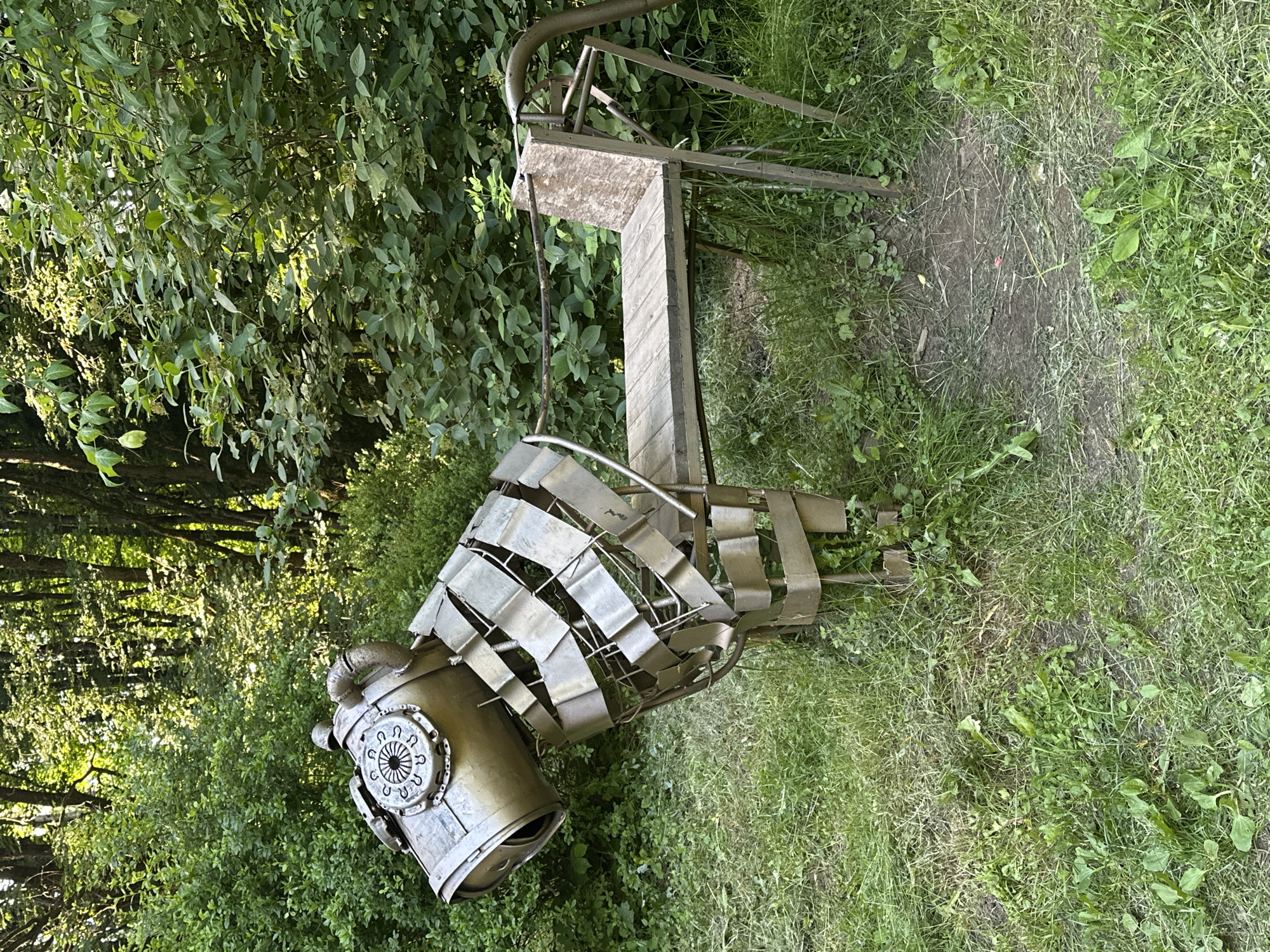 The image depicts an outdoor scene, likely a park or garden, with lush green foliage and grass. The primary object of interest is a metal sculpture that appears to be a stylized representation of an animal, possibly a horse or dog, due to its head and body structure. The sculpture is made of various metal components, including cylindrical pieces forming the head, and a segmented, cage-like structure for the body. The head has a circular detail with radiating lines, resembling a gear or a stylized eye.
In the background, there is a wooden and metal structure that looks like a bench or a seating area, integrated into the natural environment. The lighting suggests it is daytime, with sunlight filtering through the leaves of the trees, creating dappled shadows and highlights. The weather appears to be pleasant, as indicated by the vibrant greenery. No people are visible in the image. The overall atmosphere is serene and artistic, blending the man-made sculpture with the natural landscape. There is no visible text in the image.
