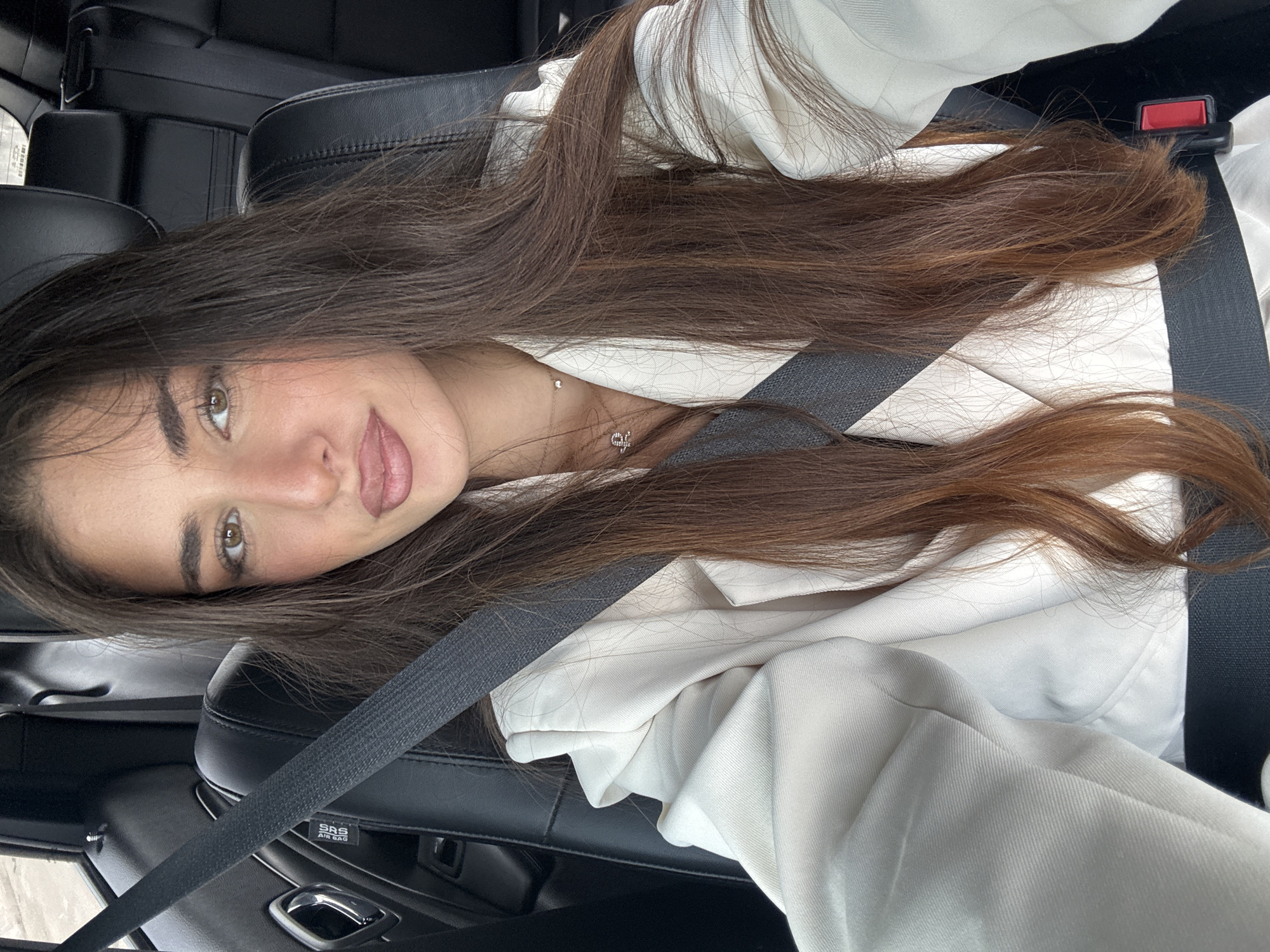 A young woman is seated in a car, wearing a white jacket, and secured with a seatbelt. She has long, brown hair and is looking at the camera. The interior of the car consists of black leather seats and dark interior panels. The location is unknown and is not North Miami Beach.