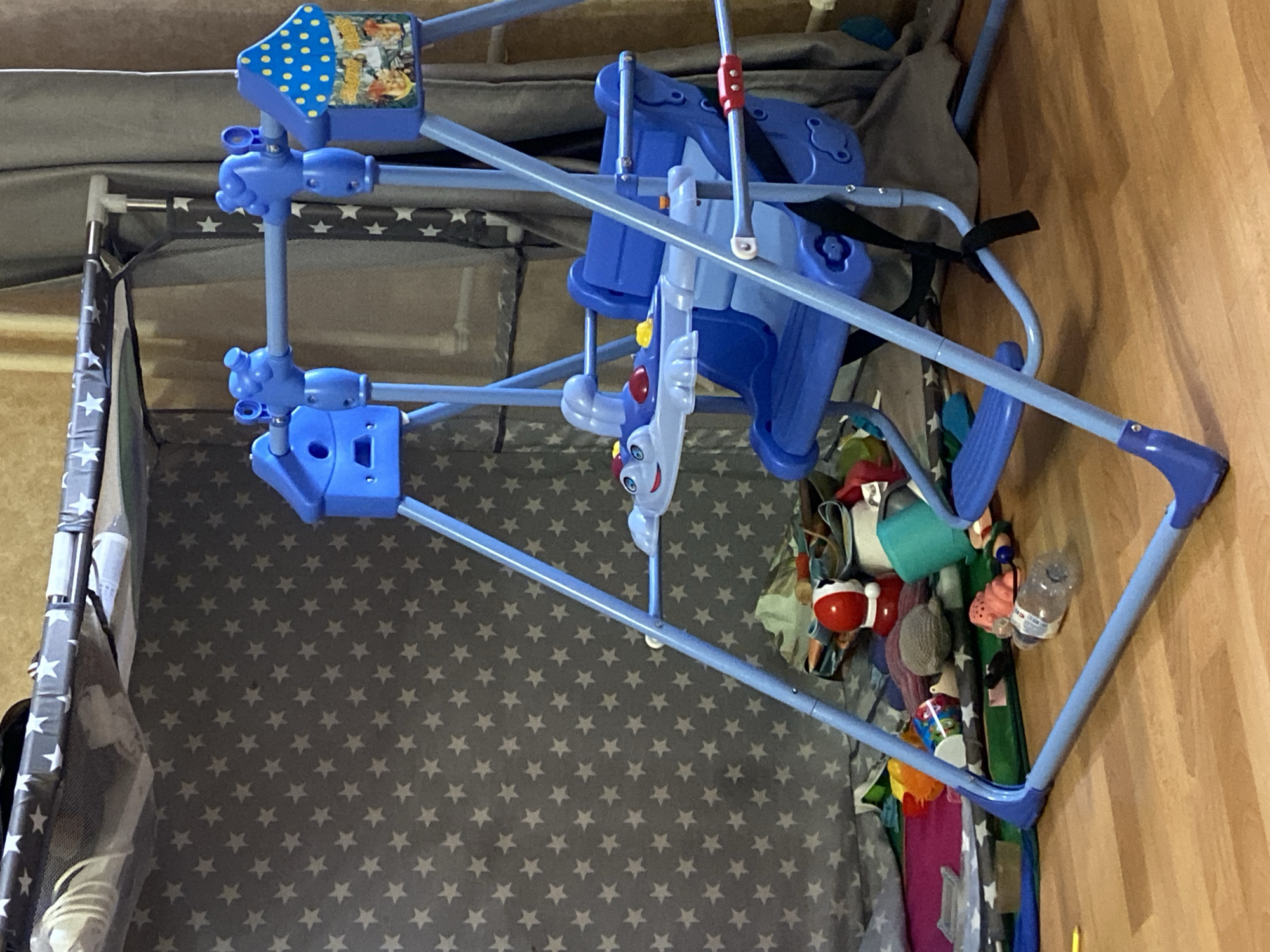 The image depicts a baby swing set suspended above a playpen. The swing is primarily blue, with hanging toys and a safety harness. The playpen has a gray and white star pattern interior. The scene appears to be indoors, with a wooden floor visible on the right side of the image. Additional items, possibly toys or baby supplies, are situated to the right of the playpen. There is no specific evidence of the location being Kazan, Russia.