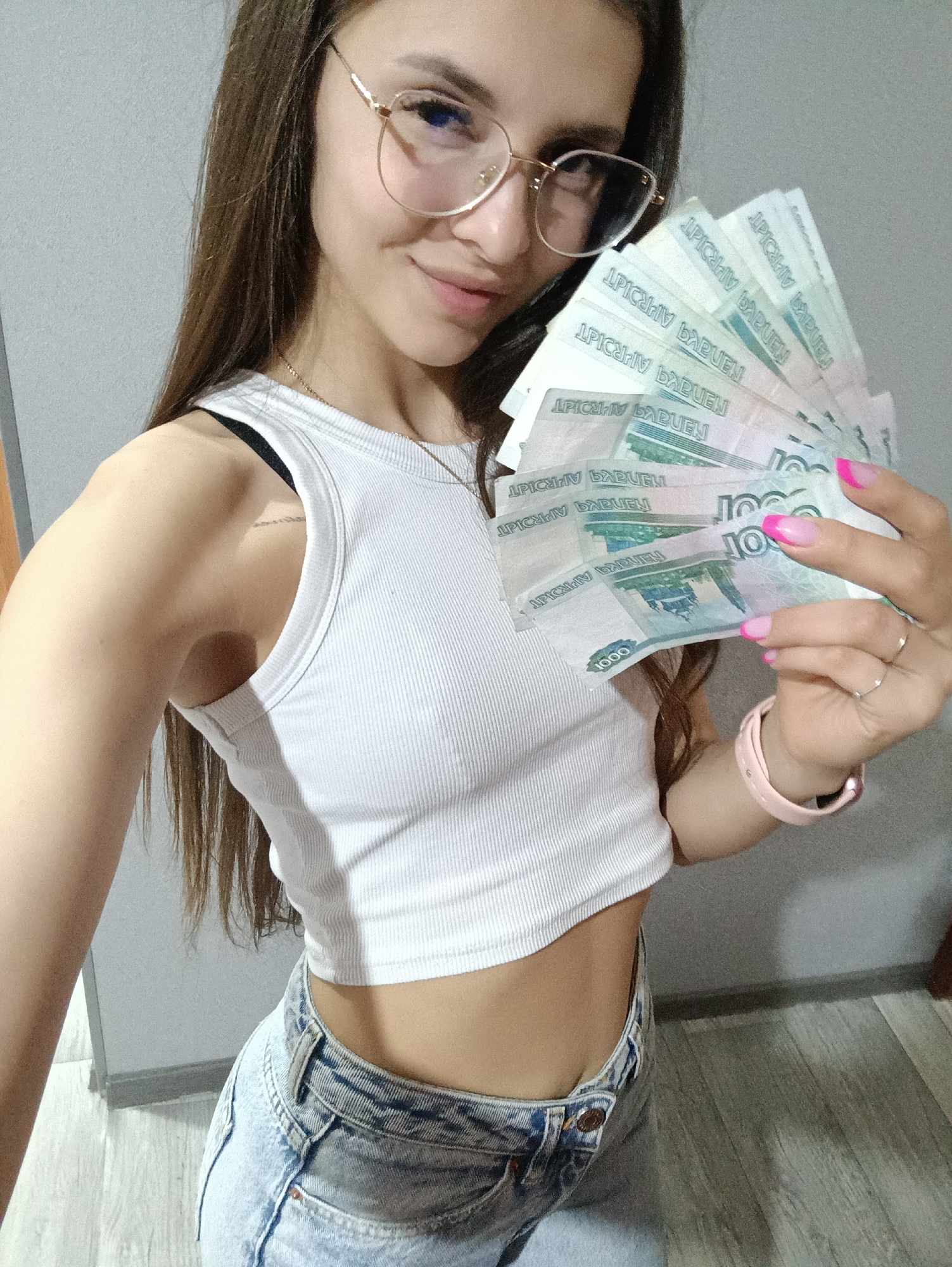 In this medium shot, a young woman wearing a white ribbed tank top, light-wash denim shorts, and glasses is holding a fan of Russian currency. The bills appear to be 1000 ruble notes. She has long brown hair and is smiling at the camera. Her nails are painted with a pink French manicure. She is also wearing a pink watch on her left wrist and two rings on her right hand. The background is a plain, light gray wall. The overall scene suggests an indoor environment.