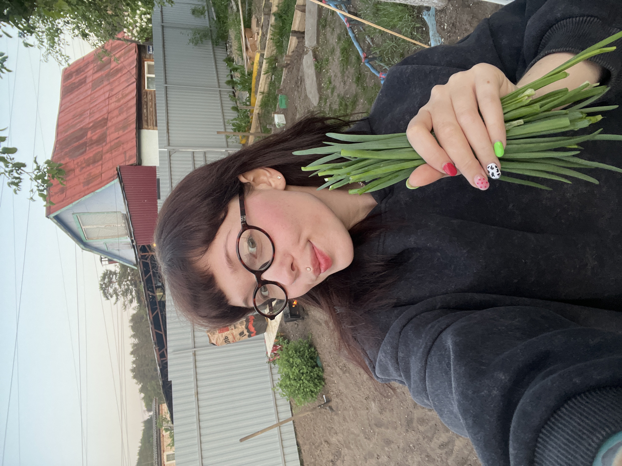 A selfie taken by a woman with brown hair and black glasses holding a bunch of green onions in her right hand. She is wearing a dark grey sweatshirt. Her nails are painted with different designs. Behind her is a garden setting with a house that has a red roof, a metal fence, and various gardening tools. The sky is visible in the upper left corner. The location is Chaikovsky, Russia.