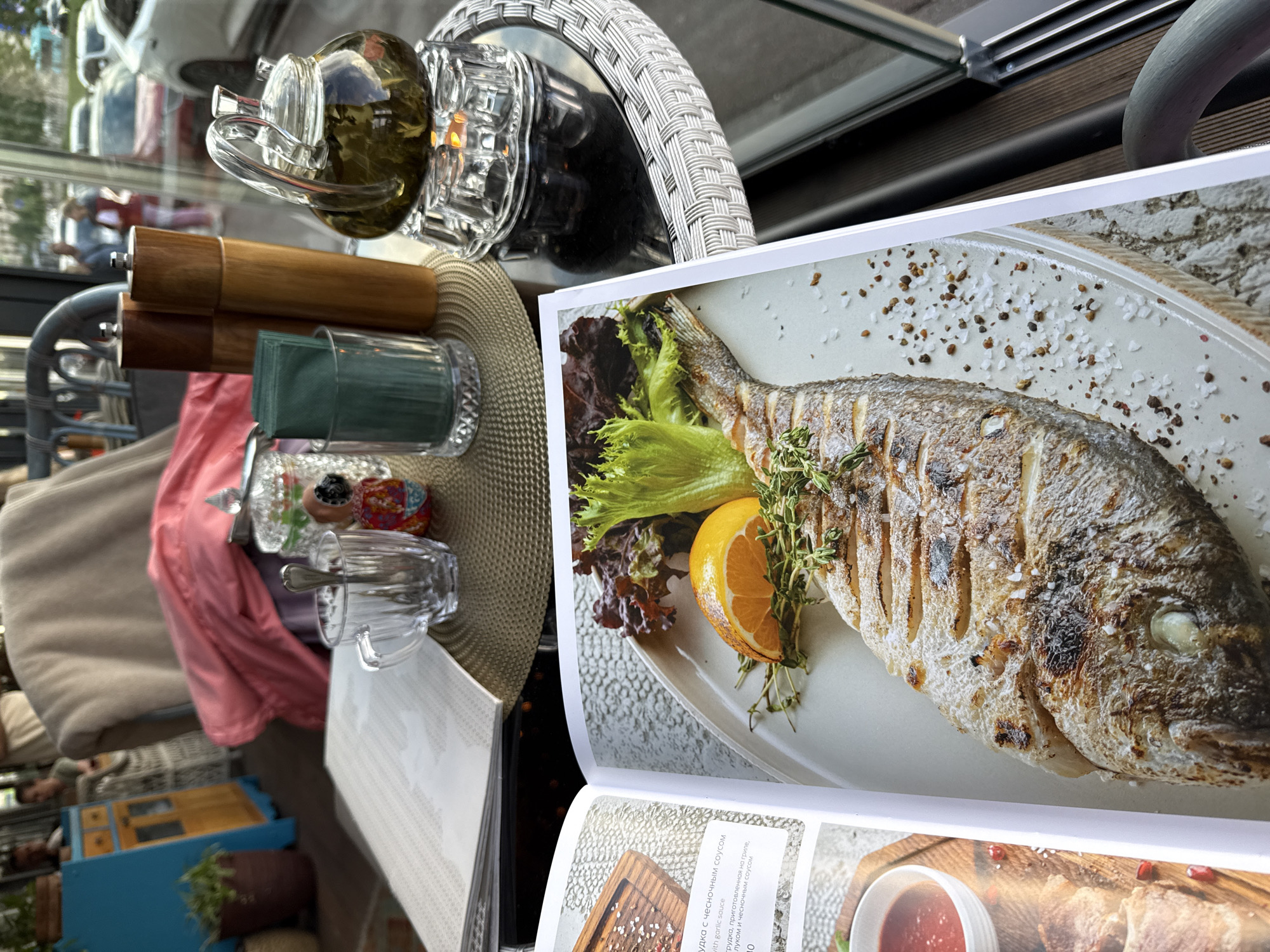 The image shows a close-up of an open menu book on a table at an outdoor cafe in Saint Petersburg, Russia. The menu displays a high-quality photograph of a whole grilled fish served with an orange slice and herbs, alongside accompanying text in Russian. The menu is placed on a woven placemat next to a transparent glass cup containing a spoon and a small, colorful figurine. Behind the cup, a glass of green tea sits on a decorative glass stand over a heat source, with two wooden spice grinders and a glass holder with green napkins to its left. In the background, out of focus, a red blanket is draped over a chair, and people are visible through a large window. The cafe setting appears to be near a street with cars passing by.