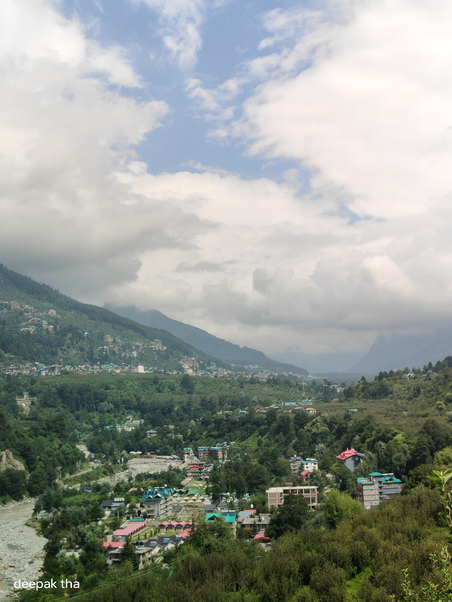 The image shows a wide, panoramic view of the town of Kalath, India, nestled in a valley between verdant, tree-covered mountains. The sky above is a mixture of blue patches and dramatic, heavy clouds. A riverbed, filled with grey stones and some water, is visible on the left side of the frame, flowing past the town. Buildings, characterized by their colorful roofs (green, red, and blue), are scattered across the slopes of the mountains and in the valley floor, indicating a densely populated area. The architecture appears to be a mix of traditional and modern styles. The overall impression is one of a scenic and developed mountain community. The text "deepak tha" is superimposed on the bottom left corner of the image.