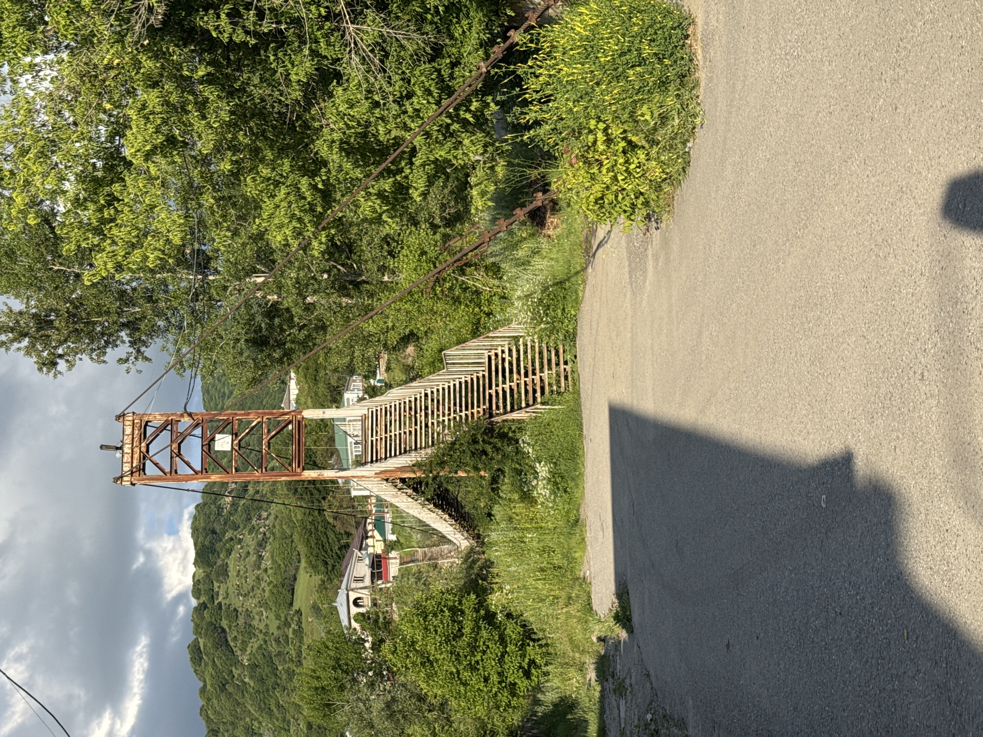 The image shows a pedestrian suspension bridge with a tall, rust-colored metal support tower. The bridge has a metal staircase leading up to it. The surrounding environment is lush and green, with trees and vegetation. In the background, there are hills and some houses. A paved road leads up to the bridge. The scene appears to be a sunny day, with some clouds in the sky. This is a view of the footbridge over the Kuban River in Karachayevsk, Russia.