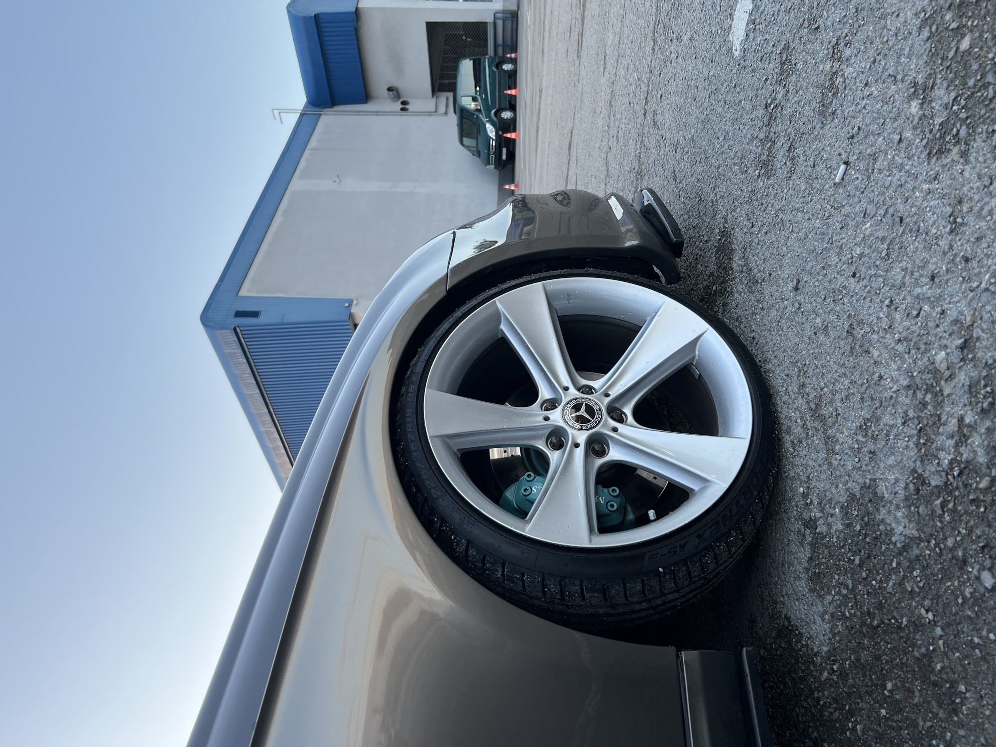 The image, oriented correctly, captures a low-angle view of the front left side of a parked car. The main subject is a light brown or champagne-colored car, likely a Mercedes-Benz, featuring large, multi-spoke silver alloy wheels. A prominent Mercedes-Benz logo is visible on the center cap of the wheel. Distinctive teal or turquoise-colored brake calipers are visible behind the spokes, with the text "Mercedes-Benz" faintly discernible on one caliper. The black tire sidewall shows visible text including "FORTUNE FSR-5" and "225/35ZR19". A black lip spoiler is attached to the lower part of the car's front bumper.
The setting appears to be an outdoor parking area, likely in an industrial or commercial zone, under a clear, bright blue sky, suggesting daytime. In the background, there is a large building with sections of grey wall and blue corrugated metal trim or roofing. Another dark green van or SUV is partially visible parked further back, with several orange traffic cones placed near it. The ground is asphalt, showing some texture and scattered debris, including a cigarette butt near the front of the main car. No people are visible, and no specific activity is taking place beyond the stationary vehicles.