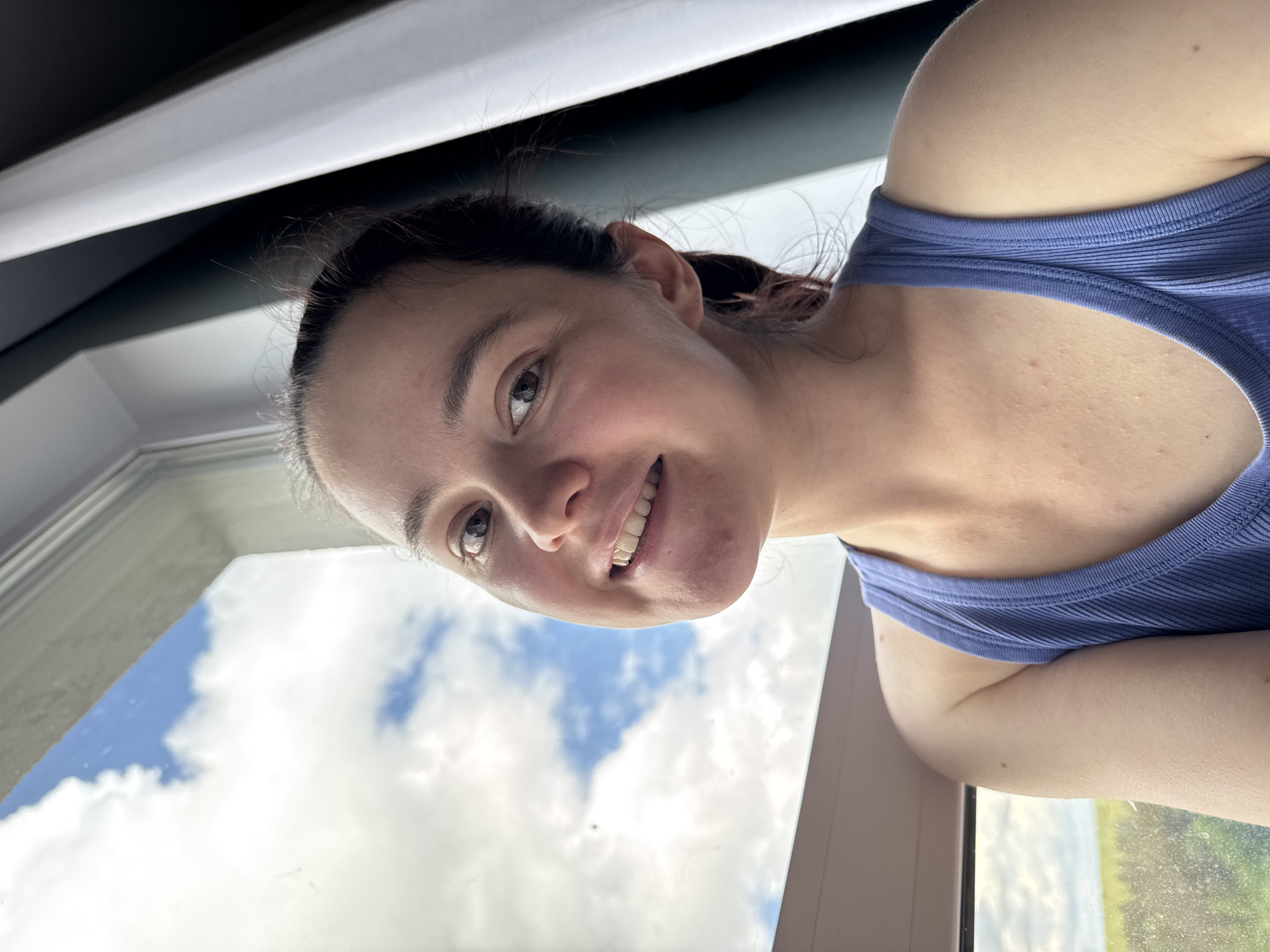 A woman with dark hair is smiling at the camera. She is wearing a blue tank top. She is next to a window with a view of clouds and blue sky. The location is not identifiable as Izhevsk, Russia based on the image.