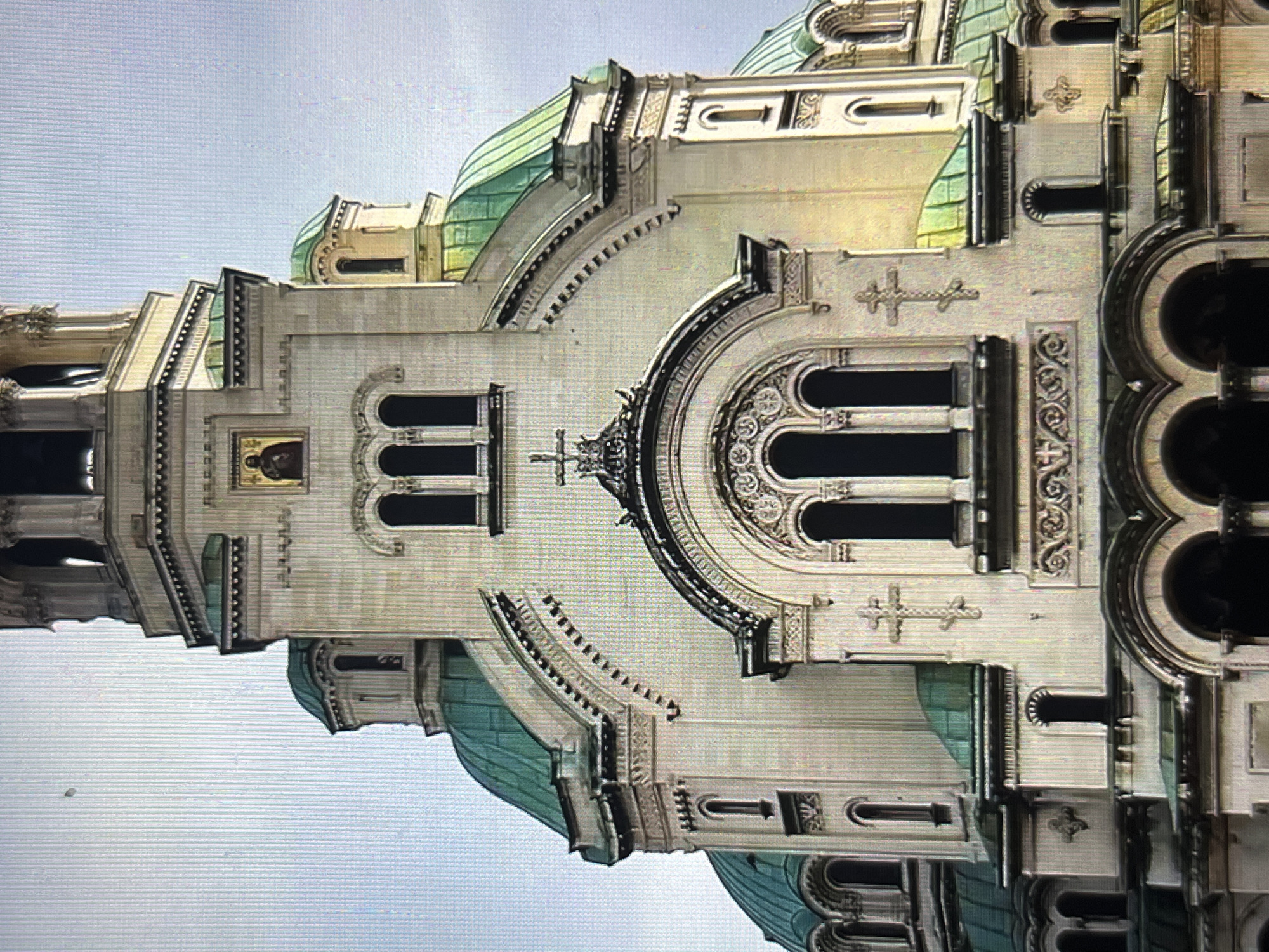 The image shows a section of a large, ornate building with pale stone or concrete walls. The architecture is characterized by arches, columns, domes covered with green tiles, and decorative elements, including crosses. Windows are dark and recessed. The style appears to be religious, possibly a cathedral or church. The sky is visible in the background, suggesting an outdoor setting. There is no sign of Tinaquillo, Venezuela, or people present in the frame.