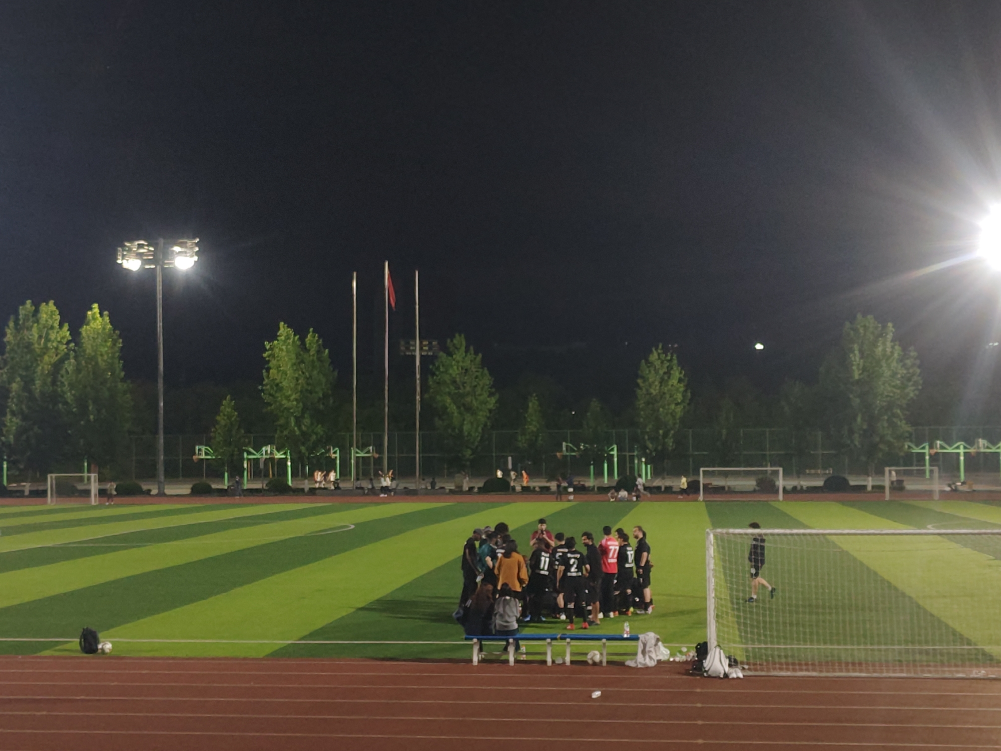 This night-time image captures an outdoor sports complex, likely in Chongqing High-tech Zone, China. The scene is dominated by a football (soccer) field featuring artificial turf with alternating light and dark green stripes, surrounded by a reddish-brown running track in the foreground.
Approximately 15-20 people, primarily men, are gathered in a tight huddle near the center of the field, suggesting a team meeting, strategy discussion, or a break during a game or practice. Many are dressed in sports attire, with some wearing jerseys displaying numbers such as 11, 2, 77, and 17. Two individuals are seated on a blue bench positioned between the running track and the football field, near the huddle. Several footballs are visible on the turf, along with a black backpack.
The entire complex is brightly illuminated by multiple tall floodlights, with a prominent light source on the right side casting a significant lens flare across the upper right portion of the image. In the background, beyond the football field, stand tall trees, fences, and various structures that appear to be other sports equipment or facilities. Two white goalposts are visible on the field, one with a net. Three flagpoles are seen in the distance, one bearing a red flag, consistent with the location context. The dark sky indicates it is evening or night, with the floodlights providing the primary illumination for the sporting activity. The atmosphere is one of active sports engagement.