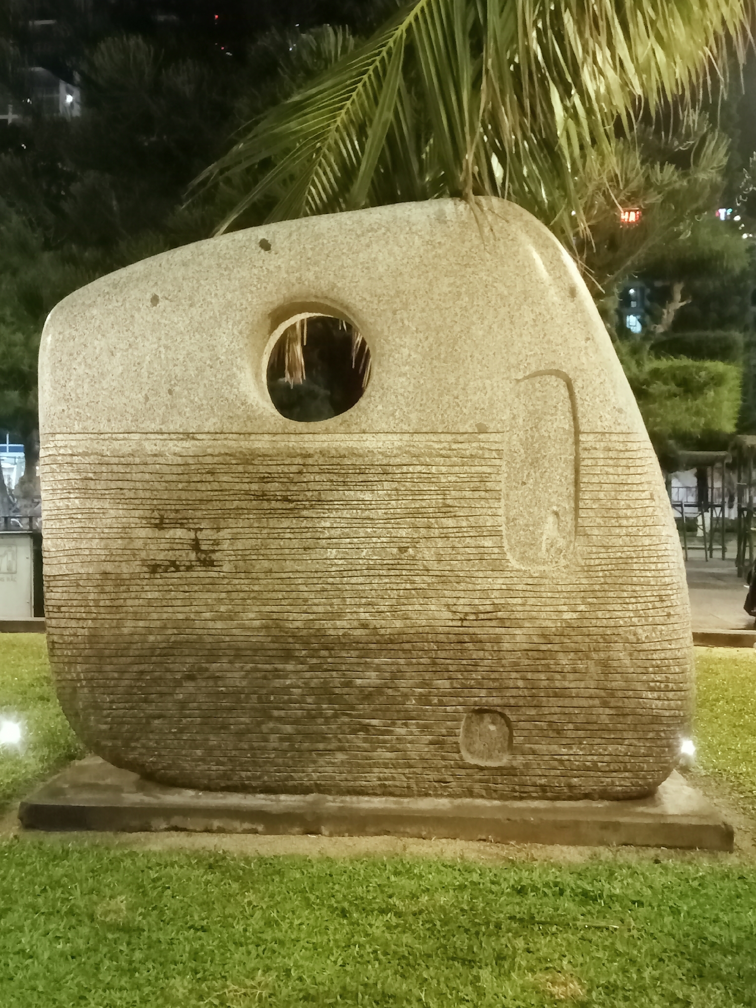 The image shows a large, grey stone sculpture in a grassy park area. The sculpture is roughly rectangular with rounded corners, featuring a circular hole near the top and horizontal lines running across the majority of its surface. There is also a vertical rectangular indentation on the right side of the sculpture, and a smaller, roundish indentation near the bottom. The sculpture sits on a concrete base. The park is illuminated with lights, and trees are visible in the background, including a palm tree. The location is in the city of Nha Trang Ward, Vietnam.