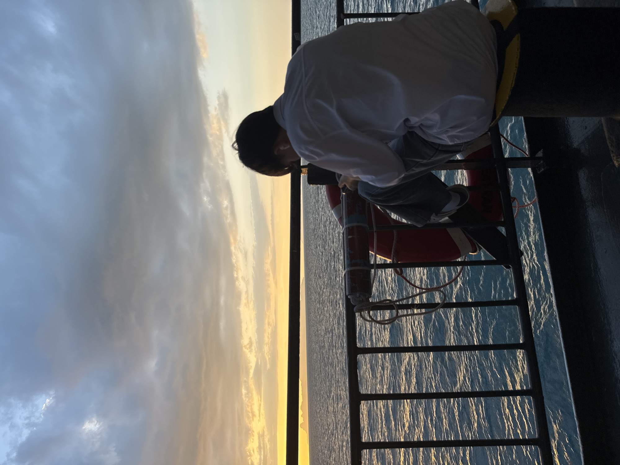 A person in a white shirt and grey pants sits on a dark bollard on a boat at sea, looking down at their phone. They are facing away from the camera, with their back to the viewer. Behind them, a metal railing separates the deck from the ocean. A red life preserver with white rope is attached to the railing. The water is a deep blue with ripples reflecting the warm tones of the sky. In the distance, a hazy landmass is visible on the horizon. The sky above is filled with dramatic clouds, transitioning from a soft yellow and orange near the horizon to a muted grey and blue overhead, suggesting either sunrise or sunset. The overall atmosphere is serene and contemplative. The location is the Andaman Sea, Thailand.