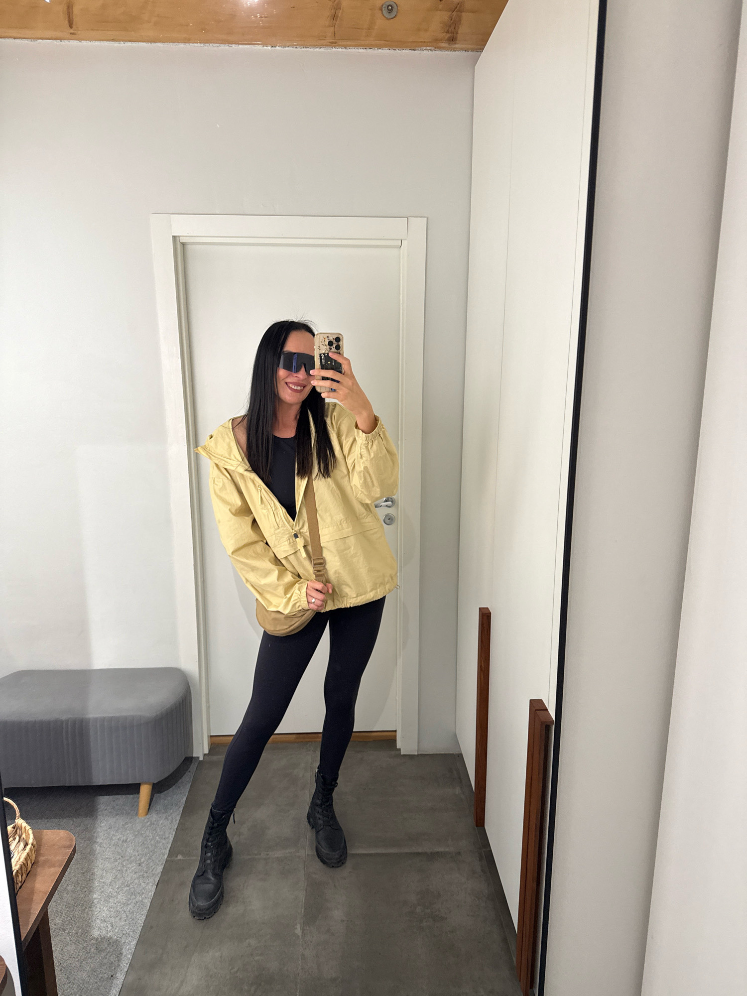 A woman stands in a room, taking a selfie in a mirror. She is wearing a light yellow jacket, a black top, black leggings, and black lace-up boots. She also has on large, dark sunglasses and a brown crossbody bag. She is smiling and has long, dark hair. The room appears to be a dressing room or fitting area, with white walls, a tiled floor, and a gray upholstered bench on the left. A wooden framed mirror is visible on the right. The image does not contain enough information to determine its location in Chaikovsky, Russia.
