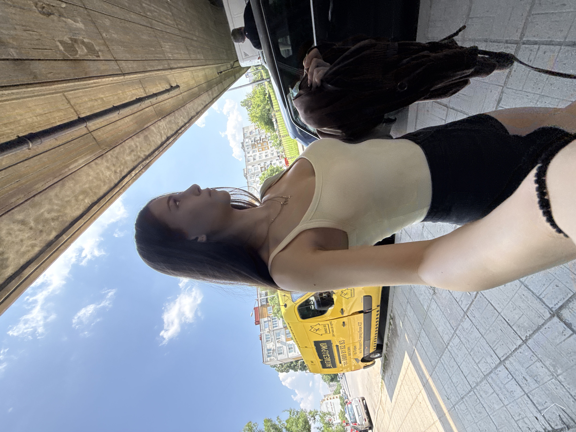 An outdoor, low-angle, eye-level shot captures a young woman standing on a tiled sidewalk, taking a selfie on a clear, sunny day in Sofia, Bulgaria. The woman, positioned on the left side of the frame, has long dark hair and is looking to her right, away from the camera. She wears a light-colored ribbed tank top, dark shorts, a delicate necklace, and an earring. Her right arm, with a black beaded bracelet, is extended towards the camera, suggesting she is holding the device. She holds a dark brown or black jacket in her left hand.
To the woman's left, a bright yellow commercial van is parked, displaying the Bulgarian text "ЖЕЛЕЗАРИЯ" (Hardware store), a phone number "0888 81 82 83", and the address "МАГАЗИН: гр. София ж.к. Младост 4 срещу блок 437" (Shop: city Sofia, Mladost 4 residential complex, opposite block 437). The van also features a house logo with the text "МИКО 80" and "ДОСТАВКА ДО АДРЕС" (Delivery to address). Behind the van, a street with parked vehicles, including a silver hatchback, and multi-story residential buildings are visible under a blue sky with scattered white clouds. Green foliage is present along the street.
The upper right portion of the image is dominated by the underside of a concrete overpass or bridge structure, featuring visible planks and a utility pipe. Below the overpass, a dark-colored car is parked, and a man with short grey hair, seen from behind in a dark shirt, stands near the vehicle. Part of a white van is also visible behind the dark car and the man. Bright sunlight casts defined shadows on the grey tiled sidewalk.