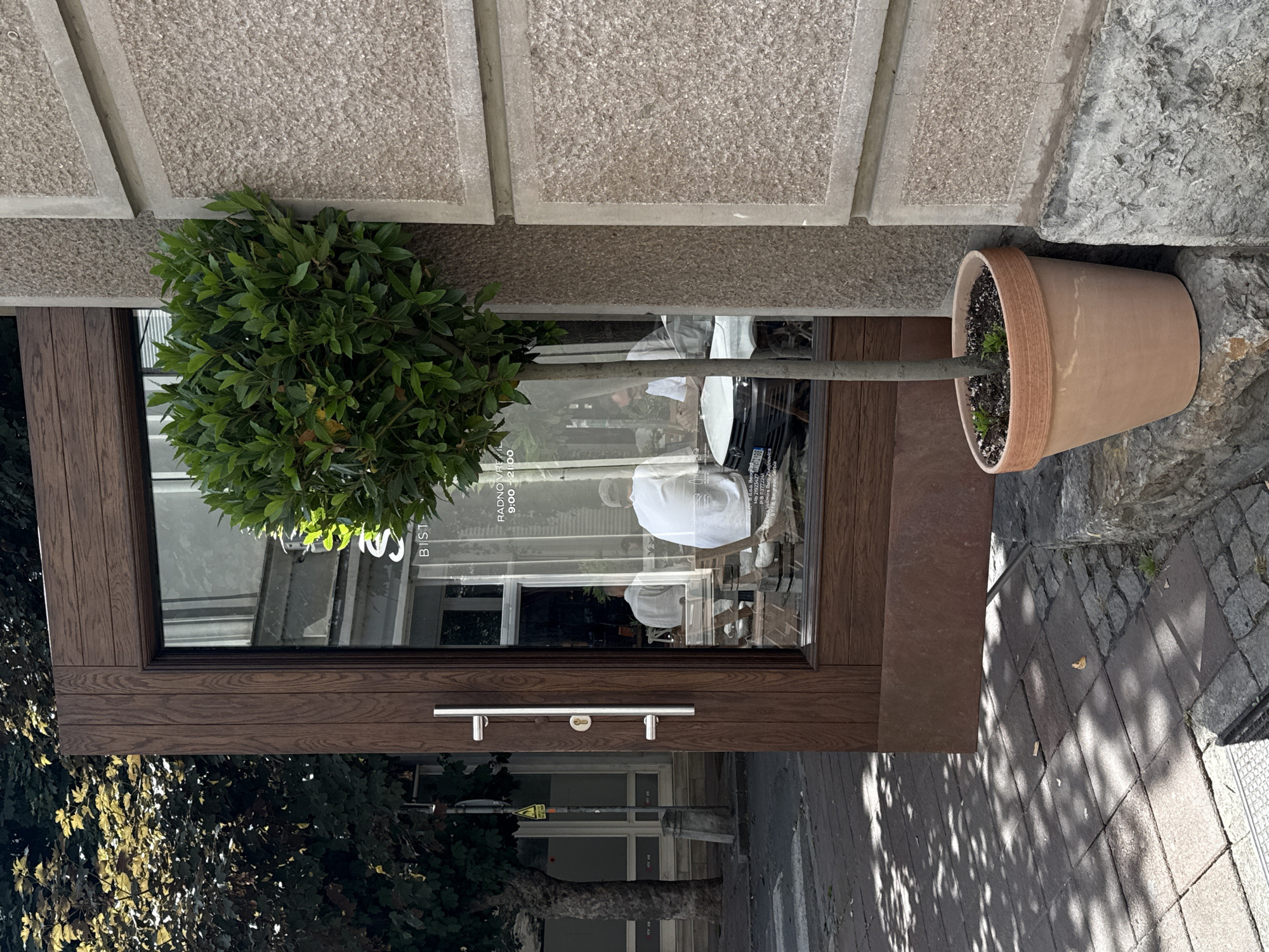 The image shows a street view of a restaurant or cafe entrance in Belgrade, Serbia. A dark brown wooden door with a large silver handle is partially open, revealing a glimpse of the interior. Inside, several people are seated at tables, and reflections on the glass show buildings across the street. A young green tree in a terracotta pot stands outside the entrance on a stone ledge, casting shadows on the paved sidewalk. The building's facade is made of rough textured stone with concrete frames. The overall scene depicts a bright, sunny day.