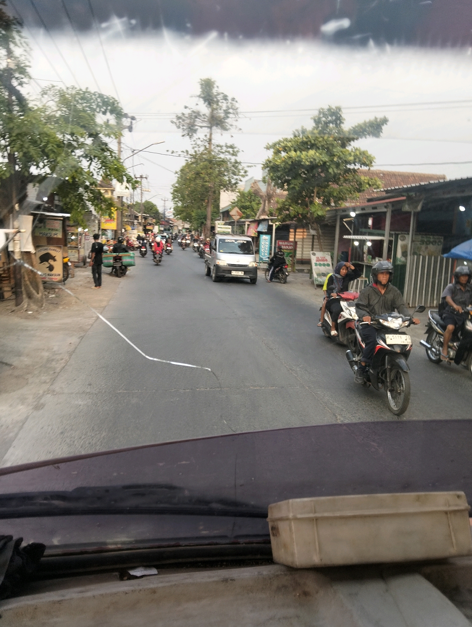 The image shows a street scene in Semarang, Indonesia, likely in the afternoon due to the lighting. The perspective is from inside a vehicle, looking out through the windshield. The road is paved and appears to be a two-lane street with moderate traffic, primarily consisting of motorcycles and a few cars.
On the left side of the road, there are several small shops or stalls with awnings and some signs. A man in a dark shirt and pants is standing on the sidewalk near one of these establishments. Several trees with green foliage line the street.
The right side of the road features more shops, some with bright lighting. Several motorcycles are visible on the road, with riders wearing helmets and some carrying passengers or cargo. One motorcycle in the foreground has the license plate "H 5178 SJ". A silver van is also present in the traffic.
The sky is overcast, suggesting it may be a cloudy day or nearing dusk. The overall impression is of a busy, everyday street scene in an urban or semi-urban area of Semarang.