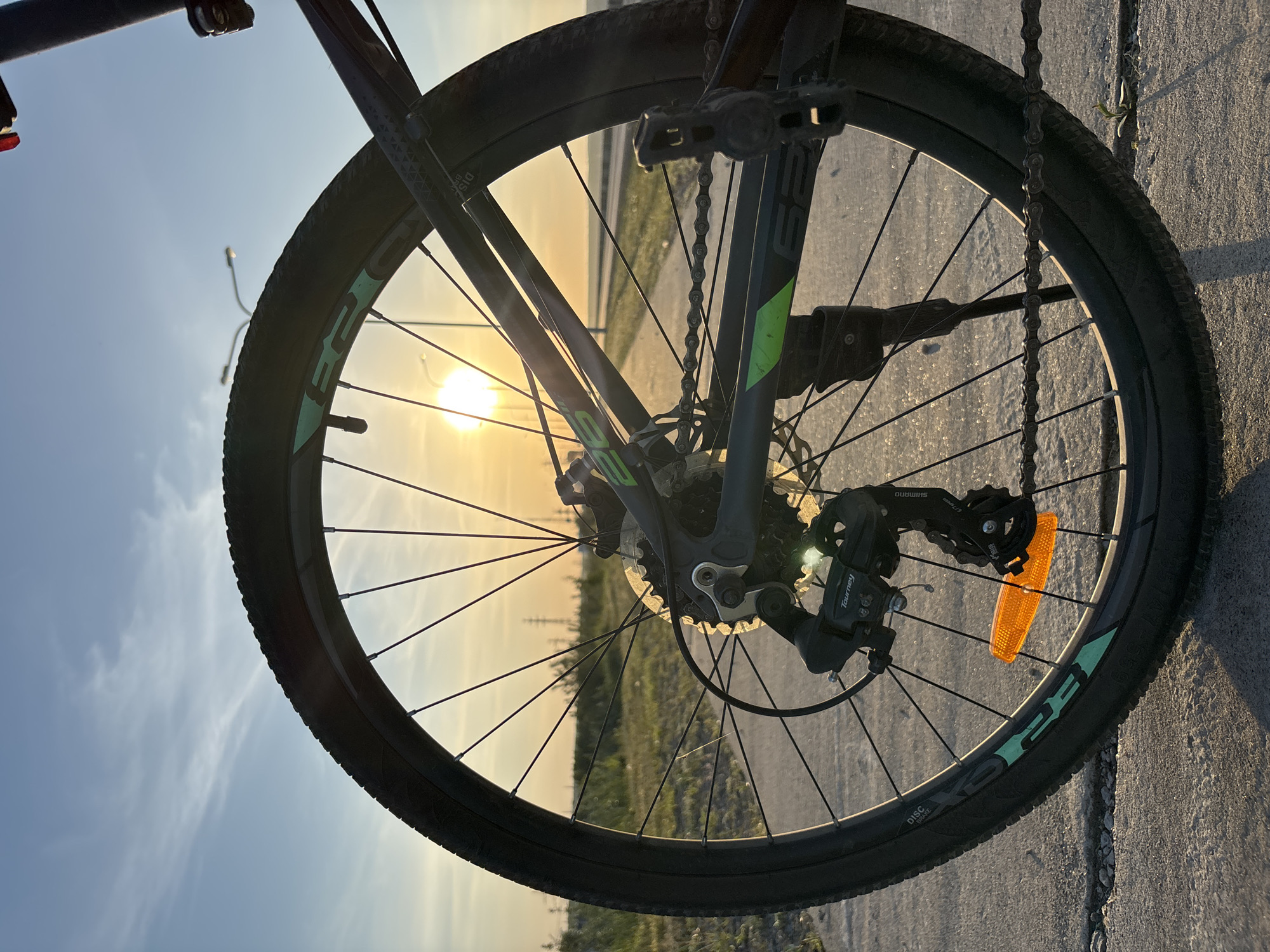 A low-angle, eye-level, close-up shot shows the front wheel of a mountain bike against a sunset or sunrise backdrop. The wheel, featuring a black tire and green lettering, is the main subject. The spokes are visible, radiating from the hub towards the tire. Parts of the bike's frame and gears are visible. The sun is positioned in the background, creating a silhouette effect. The ground is concrete with cracks and the sky fades from blue to yellow due to the sunlight. The precise location in Chaikovsky, Russia is not deducible from the image alone.