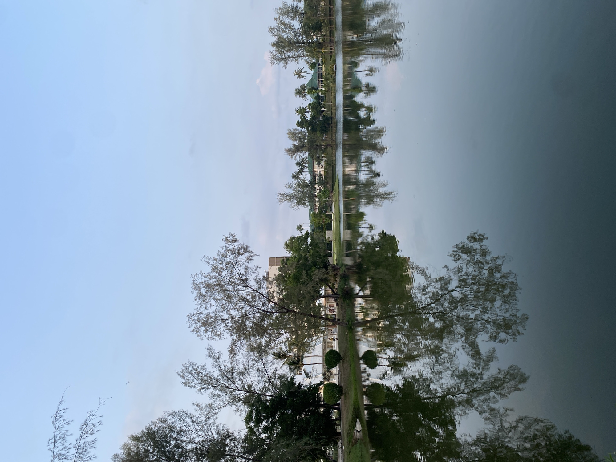 The image depicts a tranquil scene in Choeng Thale, Thailand, featuring a still body of water reflecting the surrounding environment. The reflection is clear, creating a mirrored effect of the trees and buildings lining the opposite shore. The sky is overcast, casting a soft light across the landscape. In the foreground, various trees and shrubs are visible, while in the background, buildings are interspersed among the trees. The overall impression is one of serenity and natural beauty within an urban setting.