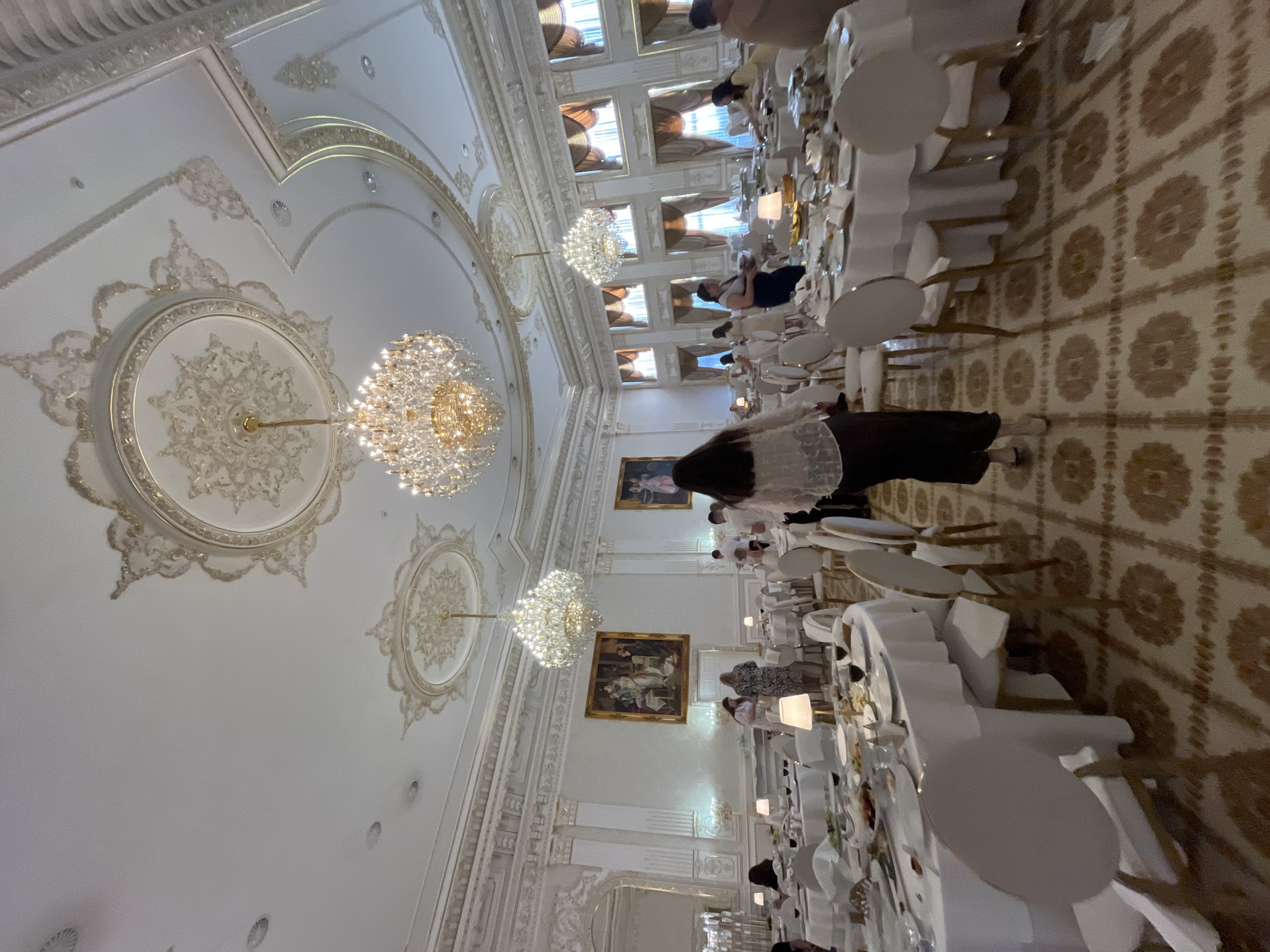 This image depicts an indoor event, likely a formal gathering or celebration, taking place in a grandly decorated hall in Dubivtsi, Ukraine. The scene is characterized by opulent decor, including ornate ceilings with elaborate plasterwork and large, sparkling crystal chandeliers. Long banquet tables are set with white tablecloths, plates, and cutlery, suggesting a meal or reception is underway.
Several people are visible, though their faces are not clearly discernible due to the angle and focus. They appear to be seated at the tables or walking through the hall, dressed in formal attire. One individual in the foreground is wearing a dark skirt and a white, lacy top. The overall atmosphere appears elegant and celebratory, with the soft lighting from the chandeliers contributing to a refined ambiance.
Notable details include the intricate patterns on the ceiling and the repetition of circular motifs. The patterned carpet on the floor also adds to the richness of the setting. The presence of framed artwork on the walls suggests the location may be a ballroom, a historic venue, or a very elaborately decorated restaurant. There is no visible text in the image. The time of day is likely evening, given the interior lighting and the absence of natural light from windows, though some windows are visible, covered by drapery, which could suggest a daytime event with controlled lighting.