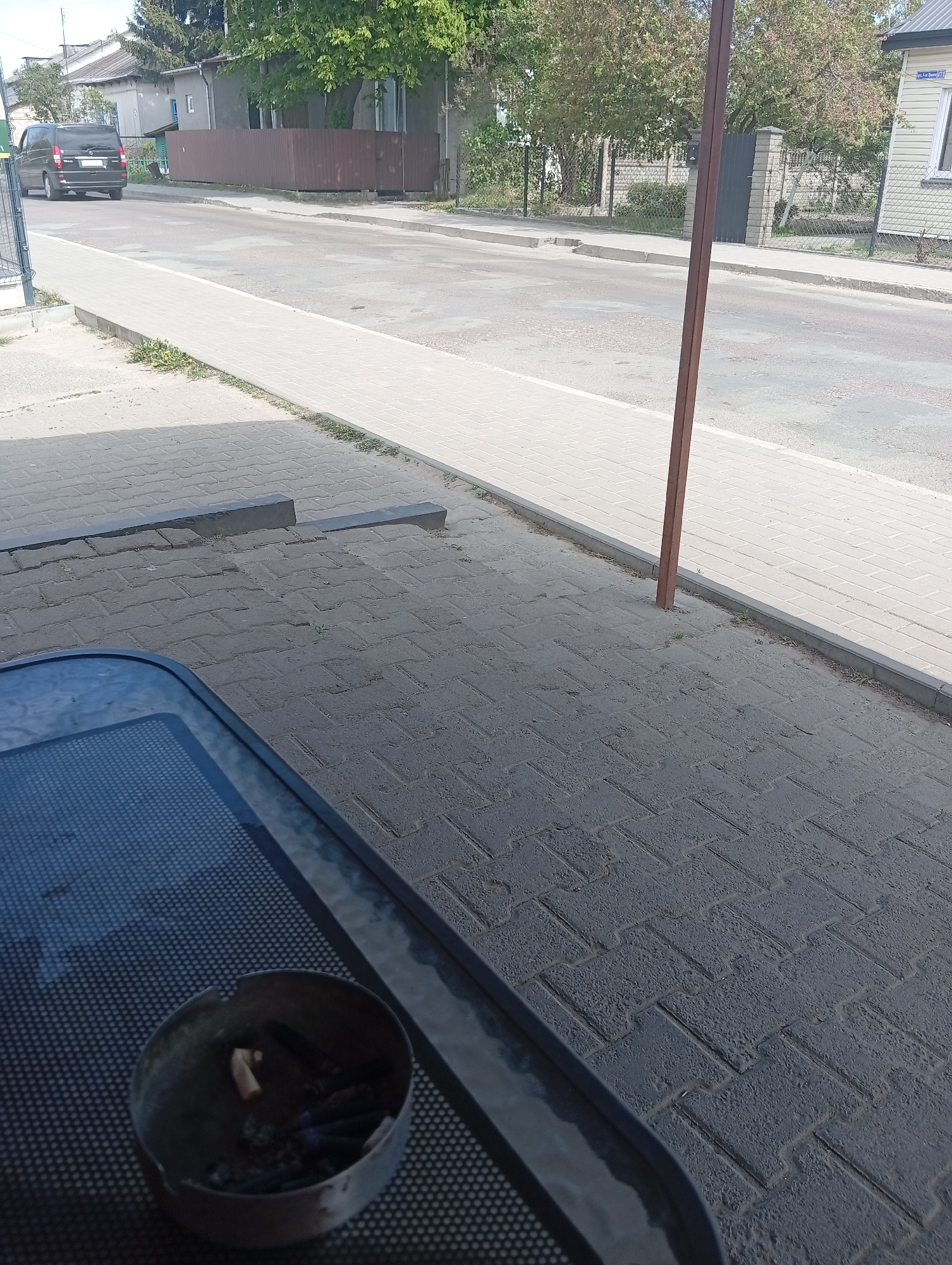 The image shows an outdoor scene in Kostopil, Ukraine. In the foreground, there is a metal ashtray filled with cigarette butts on a black metal table. The table surface has a mesh design. The ground is paved with grey rectangular bricks. A reddish-brown metal pole stands upright near the edge of the sidewalk. A paved sidewalk runs parallel to a street. On the street, a black minivan is driving away from the viewer. Houses and trees line the street. The sky is bright.