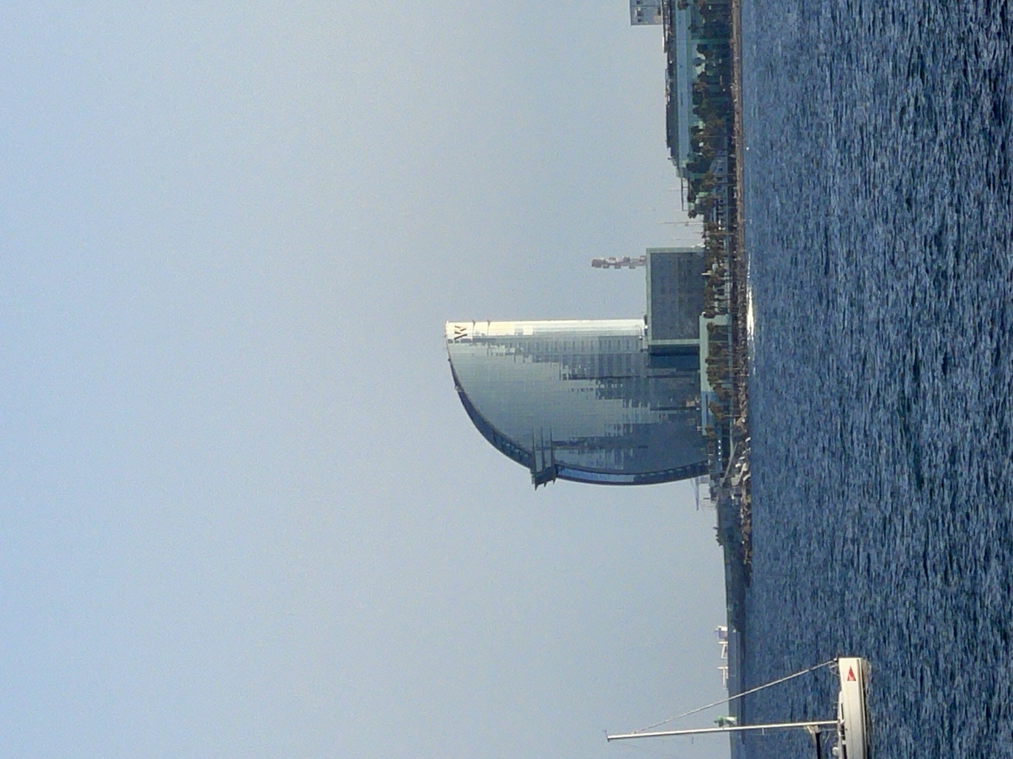 A vertical, medium shot captures the coastline of Barcelona, Spain, under a clear, pale blue sky. In the foreground, a deep blue body of water, likely the Mediterranean Sea, is rippled by a gentle breeze. To the left, a white sailboat with its mast and rigging prominently displayed is partially visible, its hull resting on the water.
Further into the scene, a distinctive curved glass skyscraper, the W Hotel Barcelona, dominates the middle ground. Its reflective facade mirrors the sky and surrounding buildings. Adjacent to it, a lower, rectilinear building complex with dark glass windows stretches along the coast, dotted with palm trees. A single red and white antenna structure rises behind these buildings. In the far distance, a faint, white lighthouse or buoy is visible on a breakwater. The overall impression is of a sunny day at the harbor.