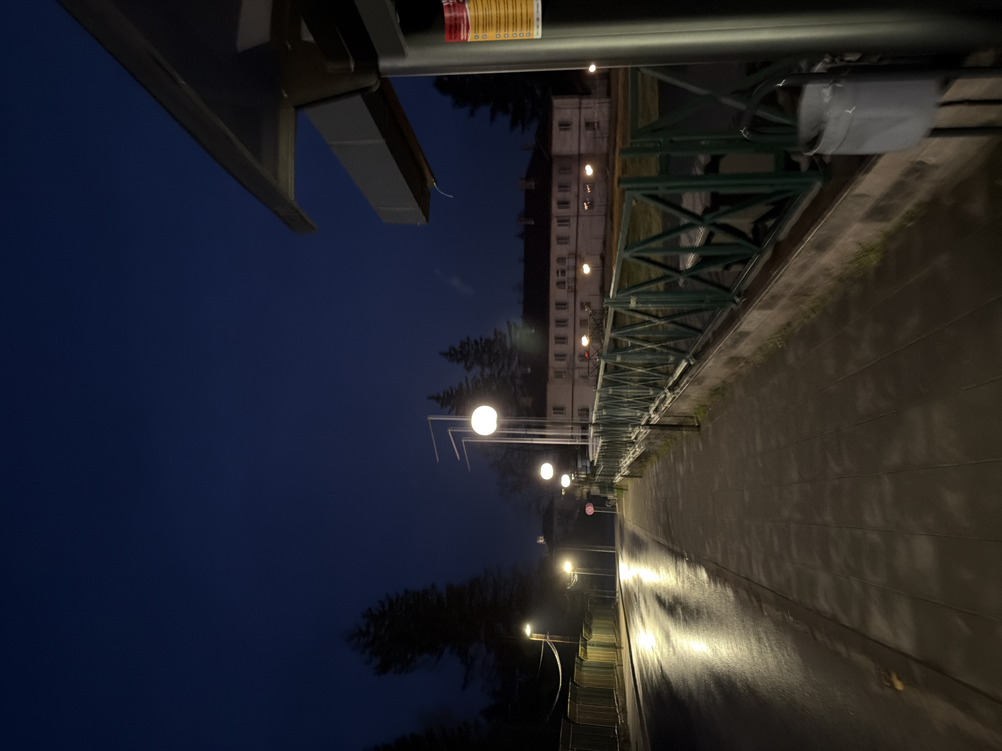 The image shows a street scene at night in Panagyurishte, Bulgaria. The view is along a walkway or road lined with streetlights and a green metal railing on one side. A building with lit windows is visible in the background. The sky is dark blue. There is an object in the upper left, a part of a metal canopy or roof structure. The surface of the walkway is wet, reflecting the light from the streetlights. There are no people visible.