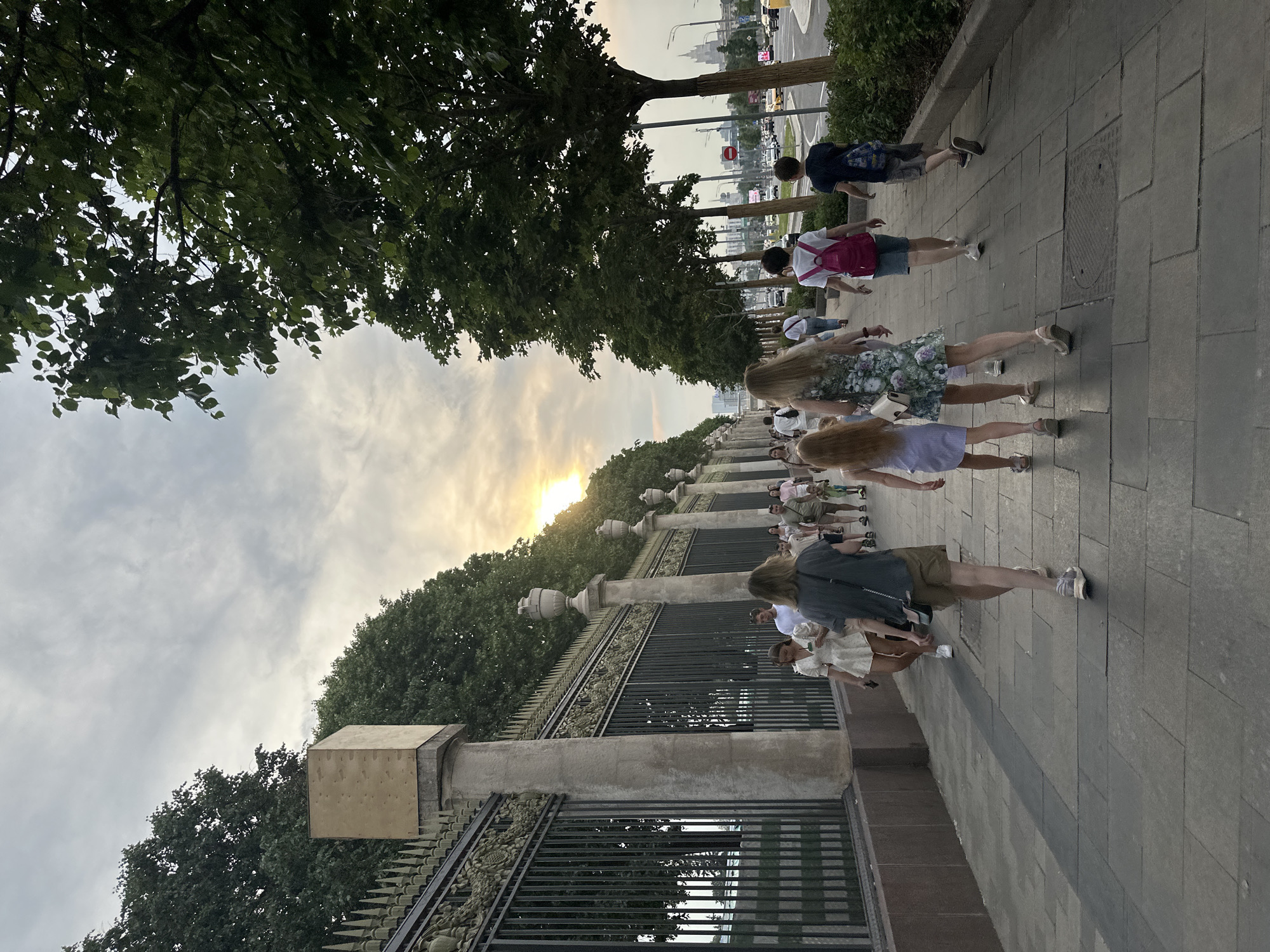 In the city of Unknown, Unknown, a group of people walks along a paved sidewalk. The sun is low in the sky, casting a warm glow and creating long shadows. The sidewalk is bordered on the left by an ornate black metal fence with stone pillars and lush green trees behind it. On the right, a row of trees lines the sidewalk, with a road and city skyline visible in the distance.
The people are walking away from the camera, dressed in casual summer clothing. Some are carrying bags, and two boys in the foreground have backpacks. The atmosphere appears relaxed, with families and individuals enjoying an evening stroll. The architectural style of the fence and pillars suggests a historic or grand area within the city.