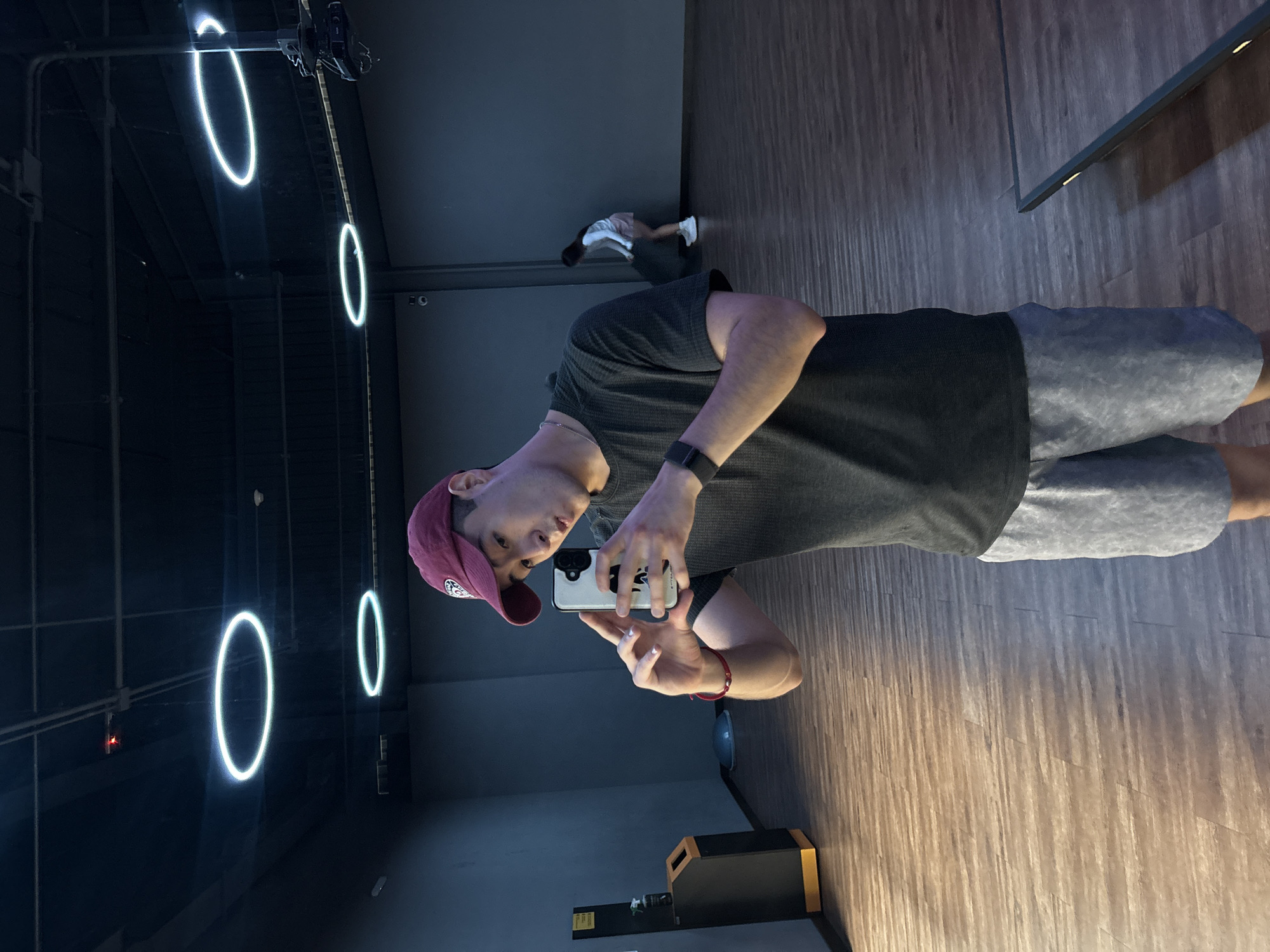 A person wearing a dark gray shirt, light gray shorts, and a maroon baseball cap is taking a selfie in a room with dark gray walls and a wooden floor. There are several neon ring lights hanging from the ceiling. A second person is faintly visible in the background, near the wall. The image was taken in an indoor space in San Bernardino Tlaxcalancingo, Mexico.