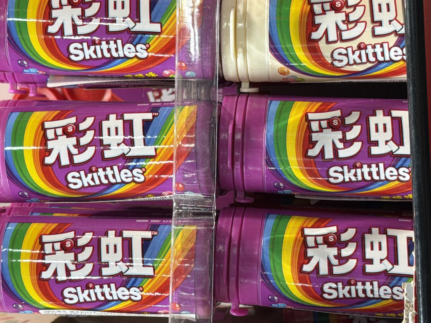 This close-up shot captures multiple cylindrical packages of Skittles candy displayed vertically in what appears to be a retail store setting in Penza, Russia. Most of the visible packages are a vibrant purple, featuring a prominent rainbow graphic on their labels. One cream-colored package is partially visible at the top right, suggesting a different flavor or variant. The packages are stacked on a shelf, likely for sale.
The primary visible text includes the English brand name "Skittles" and the Chinese characters "彩虹" (Cǎihóng), which translate to "rainbow." A "TM" (trademark) symbol is also visible. Smaller numbers like "500-4500", "830027", "8-2-07", "5064900", and "5064000" are printed on the packaging, possibly indicating product codes or dates. The bright, artificial lighting suggests an indoor retail environment.