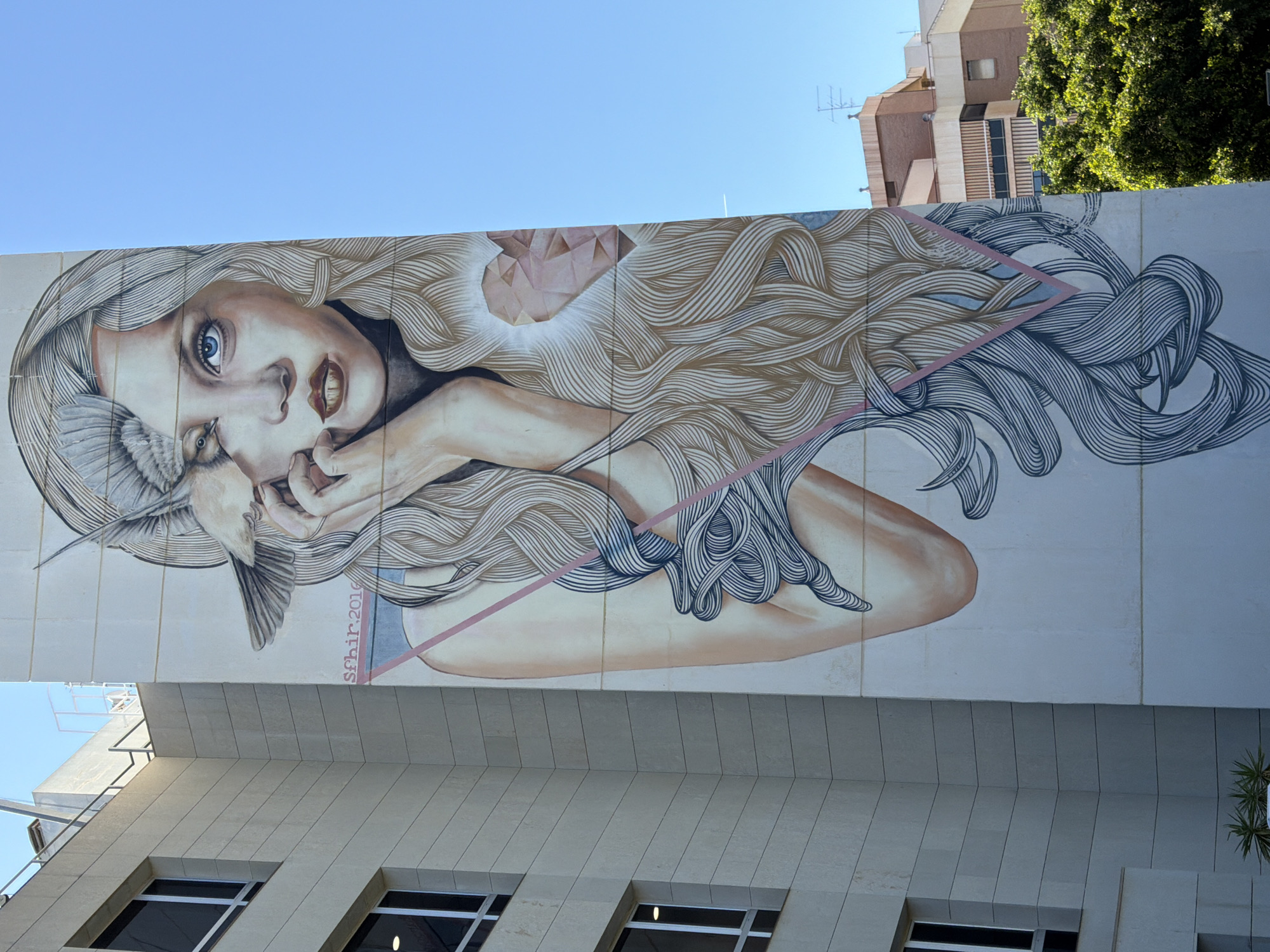 A vertical eye-level shot shows a building mural in Unknown, Unknown. The mural depicts a woman with long, wavy hair in light brown and gray hues. She has fair skin, blue eyes, and a pensive expression. She holds a bird near her face with her hands, which are tan in color.
Her hair is styled in a way that gives the illusion of movement. At the top of her hair, a geometric shape is visible. A pink line is drawn around the figure.
The building beneath the mural has a stone façade with several windows visible. Above the mural, other buildings are partially visible, as well as a tree. The sky is clear and blue.