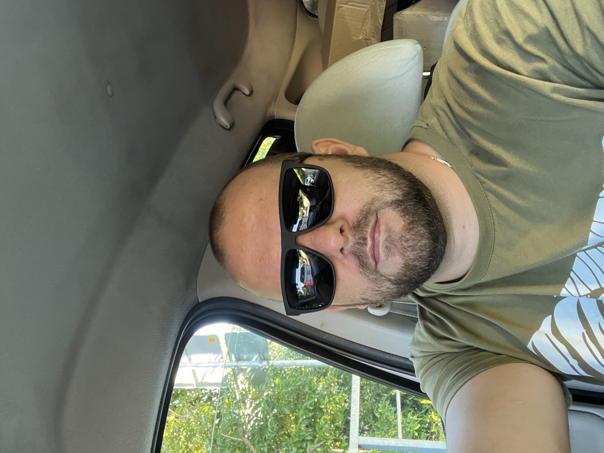 A man is depicted inside a vehicle during the daytime, likely posing for a photo. He has a beard, a bald or closely shaved head, and is wearing black rectangular sunglasses. He wears a military-green t-shirt with a large white graphic design visible on the right side, and a thin silver chain around his neck.
The interior of the vehicle, possibly a car or van, features a gray headliner, a light-colored grab handle, and a headrest behind him. The rear section of the vehicle appears partially filled with cardboard boxes. Through the side window, bright green foliage is visible, indicating a sunny outdoor environment. Reflections in his sunglasses show street traffic, including a white van and a red traffic light, suggesting the vehicle is on a road.
The scene is set in Sochi, Russia. The man's expression is neutral, largely obscured by his sunglasses. Some indistinct letters are faintly visible on a white sign outside and on a vehicle reflected in the sunglasses, but no clear text can be deciphered.