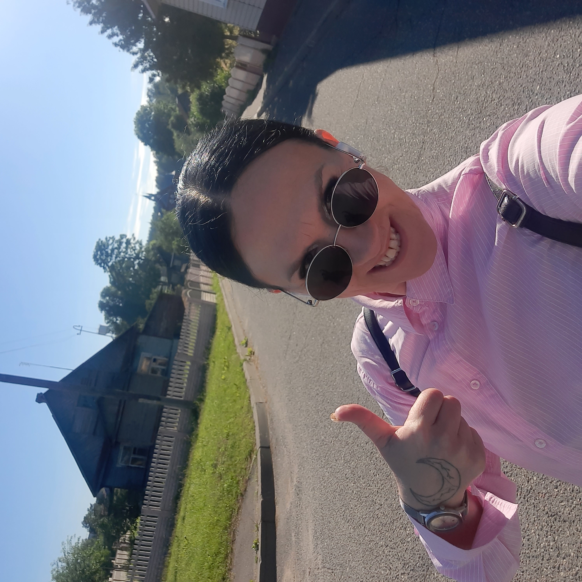 In a selfie taken outdoors, a woman stands on an asphalt road, giving a thumbs-up. She is wearing a pink striped button-down shirt and round sunglasses. A black strap from a bag is visible across her shoulder. On her left hand, she has a crescent moon tattoo. In the background, a wooden house with a fence and trees are visible under a clear blue sky. The scene appears to be in the city of Parechcha, Belarus, based on the provided context.