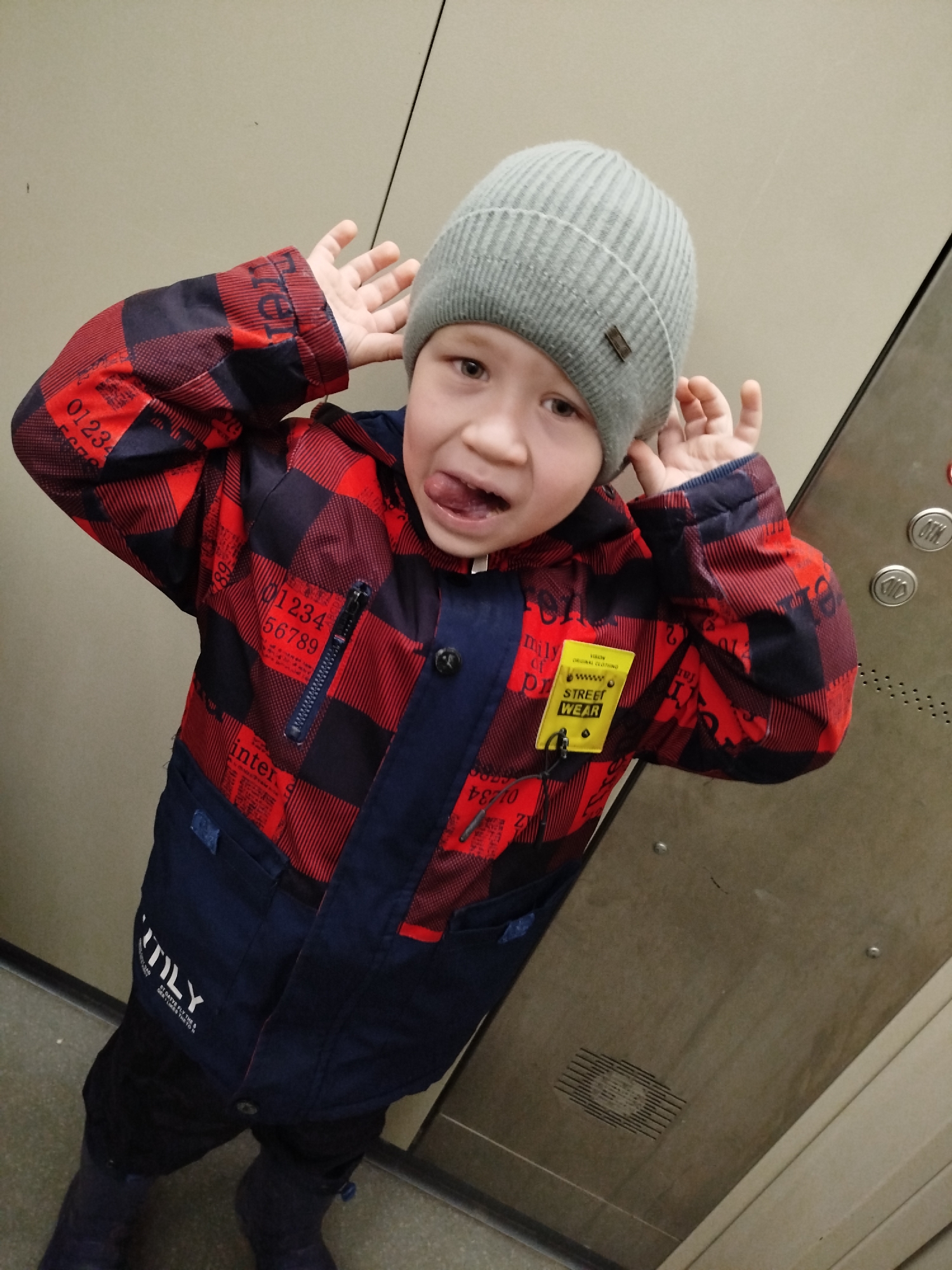 A young boy wearing a grey knit hat and a red and black checkered jacket with blue accents is standing in an elevator. He is sticking his tongue out and holding his hands to his ears, making a playful face. The elevator has metal walls and button panels. The jacket has numbers printed on it and a yellow tag that reads "STREET WEAR." The lower part of the jacket and the boy's pants and boots are dark blue. The location may be Chaikovsky, Russia, based on text on the jacket.