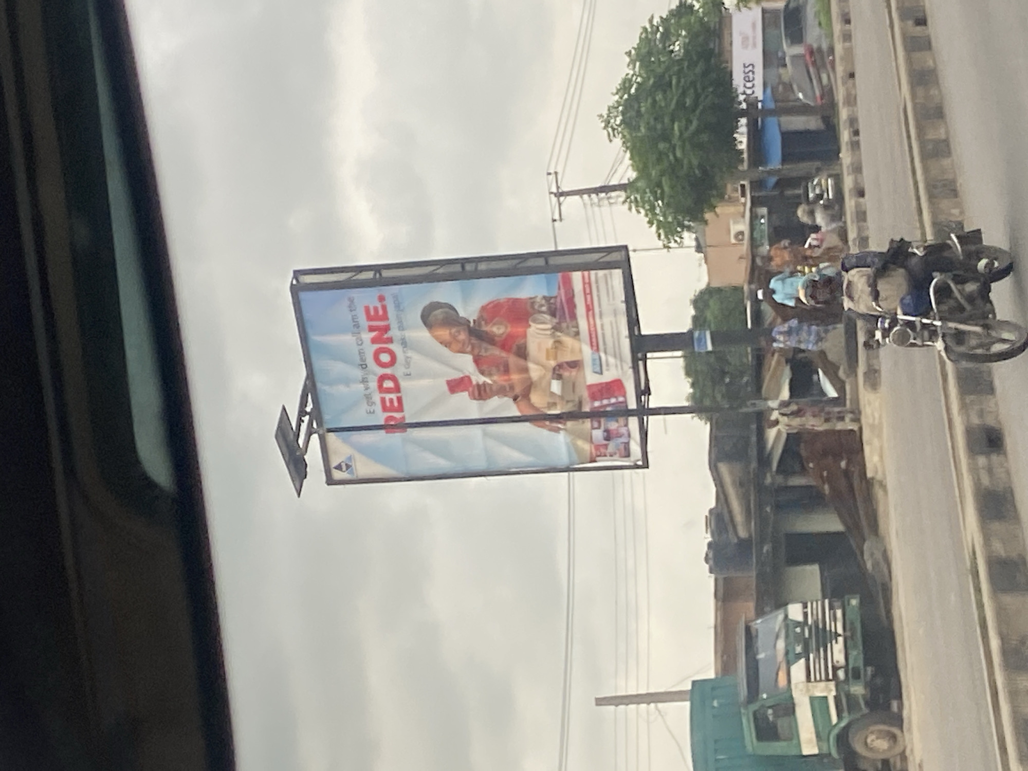 A billboard advertising "RED ONE" is positioned prominently in Ikeja, Nigeria. The billboard features a woman holding a red object and text in a local dialect. In the foreground, a motorcyclist wearing a backpack rides along a road. Further back, a green and white truck is parked near some buildings. The scene depicts a busy urban environment with people and vehicles.
