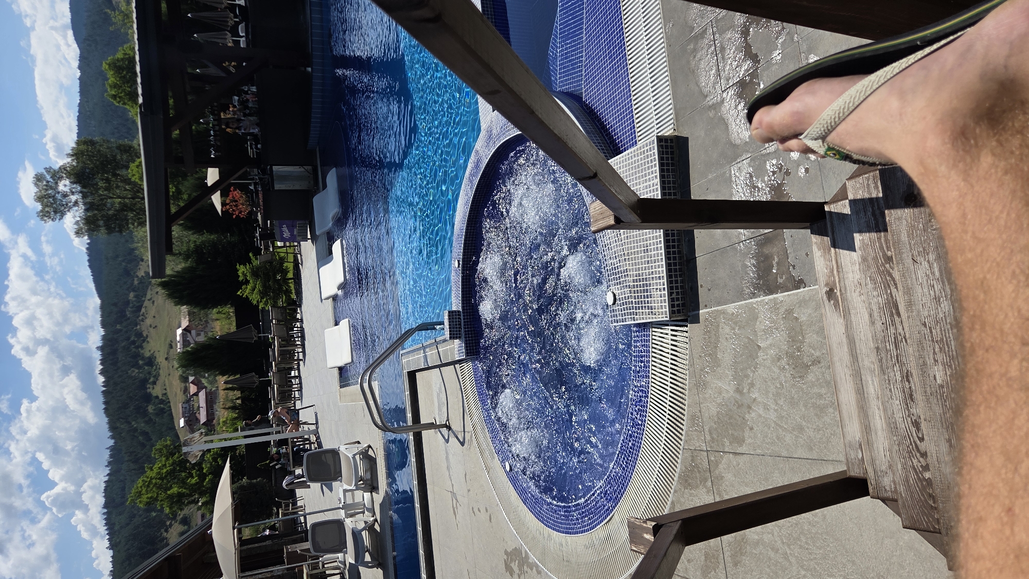 The image depicts an outdoor swimming pool area, likely at a resort or hotel, with a mountainous backdrop characteristic of Bania, Bulgaria. In the foreground, a person's foot wearing a flip-flop is visible on wooden stairs leading down to the pool. The pool itself is a deep blue, with a section featuring a bubbling hot tub tiled in blue and white mosaic. Sun loungers and tables with umbrellas are visible on the pool deck, suggesting a recreational and relaxation area. The landscape behind the pool features a dense forest covering rolling hills and scattered houses, typical of the rural mountainous scenery of Bania. The sky is partly cloudy, indicating fair weather.