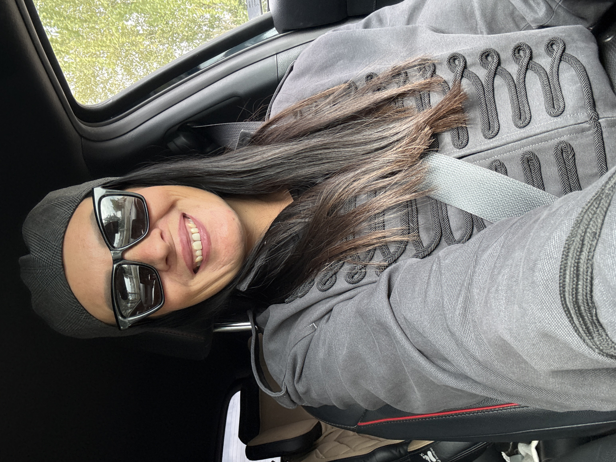 A woman wearing sunglasses and a grey beret smiles while sitting in the driver's seat of a car. She has long, dark hair and is wearing a grey jacket with black detailing. The seatbelt is fastened across her chest. The car's interior is visible, including the dashboard and a portion of the passenger seat. Outside the car, green foliage can be seen. The location of this image is unknown and cannot be determined to be Chaikovsky, Russia.