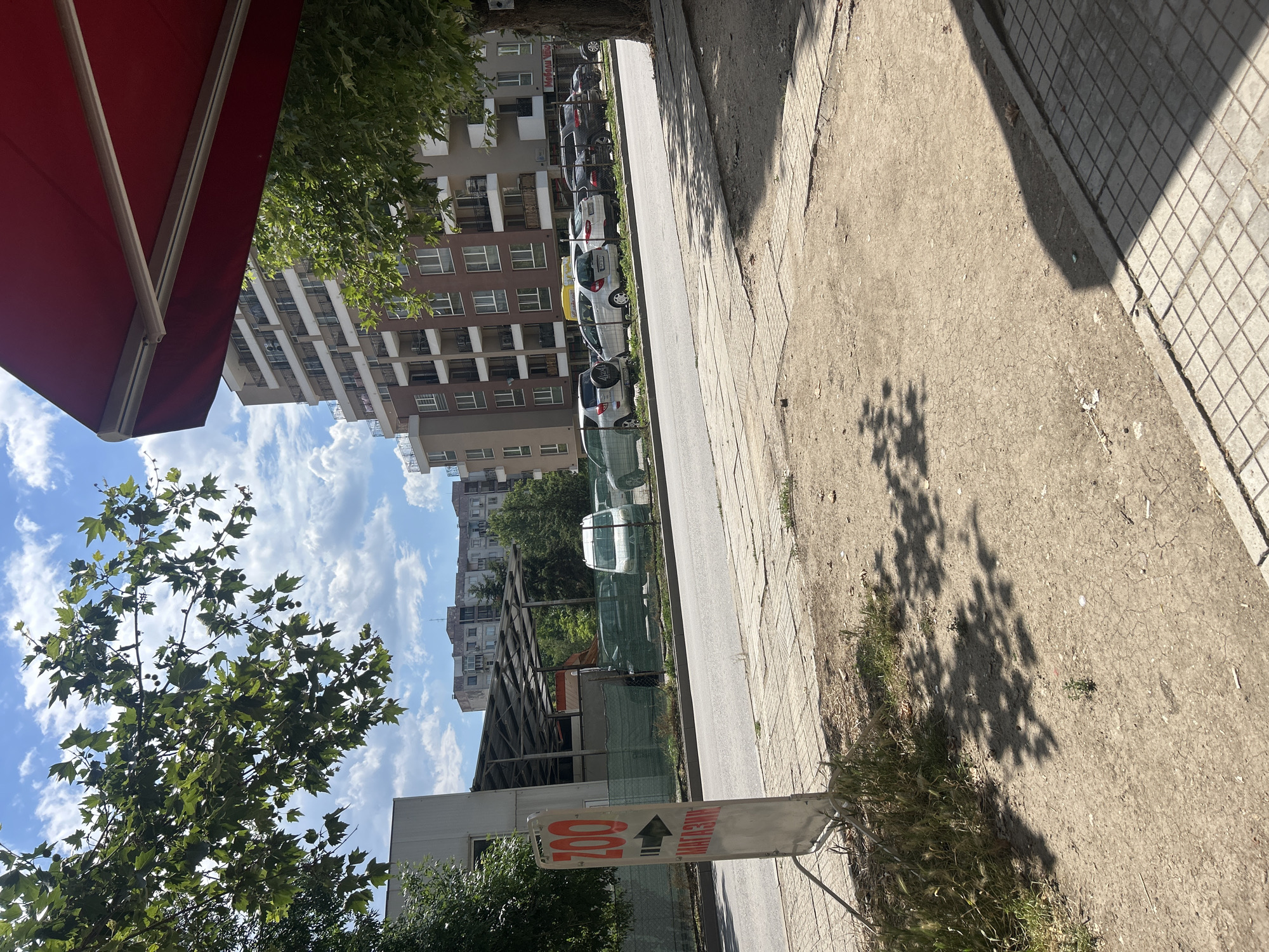 The image captures a street scene in Plovdiv, Bulgaria, on a sunny day with a partly cloudy sky. In the foreground, a paved sidewalk transitions to a dirt and gravel area with sparse vegetation and shadows cast by trees. A white, rectangular sign with red lettering is prominently displayed, indicating "ZOO" with an arrow pointing to the right. Below "ZOO," in red lettering, are the Cyrillic letters "МАГАЗИНИ," which translates to "SHOPPING."
Adjacent to the sidewalk is a paved road where several cars are parked and driving. To the left of the road, behind a green fence, is a covered structure with a metal roof supported by posts. A white van is visible parked under this structure, along with other vehicles and construction fencing. Further in the background, a multi-story residential building with many balconies and windows stands tall. Trees with green leaves are scattered throughout the scene, and a red awning is partially visible in the upper right corner. The overall impression is a typical urban street with commercial and residential elements.