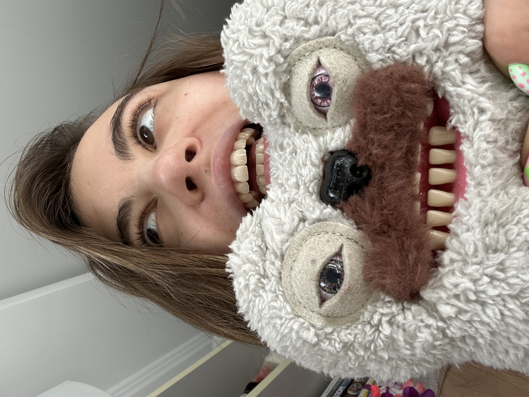 A close-up, low-angle shot shows a young woman holding a fluffy stuffed animal with a cartoonish face in front of her own. The stuffed animal has beady eyes and a wide, toothy grin. The woman's mouth is also open, revealing her teeth, and she is looking upwards and to the left. Her brown hair frames her face. The background is a plain, light-colored wall and ceiling. The stuffed animal is white with brown accents around its mouth. The woman's fingernails are visible at the bottom right of the frame, painted with a green and pink polka dot design. The setting is indoors. There is no indication of the location being Moscow, Russia.