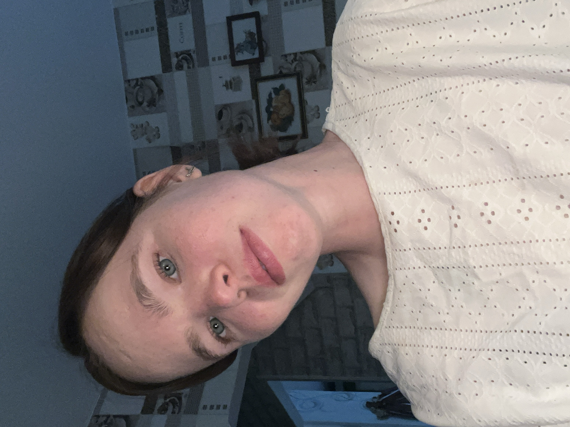 A young woman is pictured indoors, likely in a domestic setting in Chaikovsky, Russia. She is facing the camera at an oblique angle, wearing a white eyelet top. She has fair skin, light eyes, and brown hair. Behind her, the wall is covered with patterned wallpaper and two framed pictures are visible. A portion of a white table can be seen at the bottom of the frame. The lighting is dim.