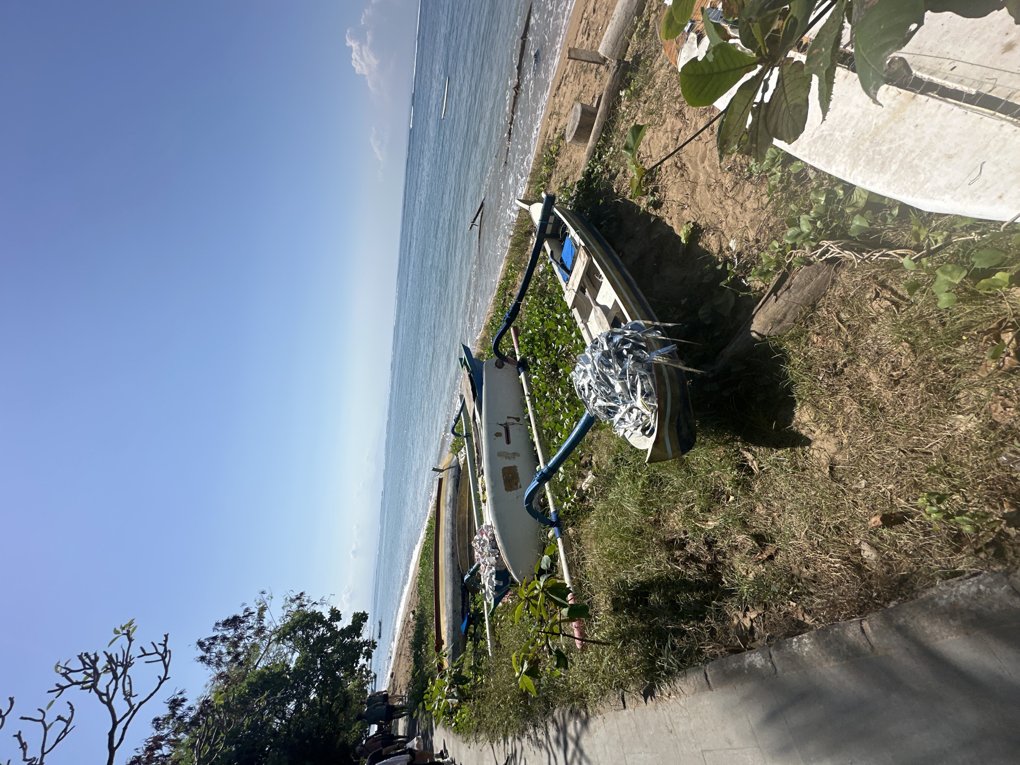 The image depicts a sunny day on a beach in Denpasar, Indonesia. Several traditional outrigger canoes are pulled up onto the sandy shore, resting on a patch of dry grass and green vines. The ocean stretches out to the horizon under a clear blue sky, with a few wispy clouds visible. To the left of the scene, a paved walkway runs along the edge of the beach, and a group of people are walking away from the camera. The overall impression is one of a tranquil coastal setting.