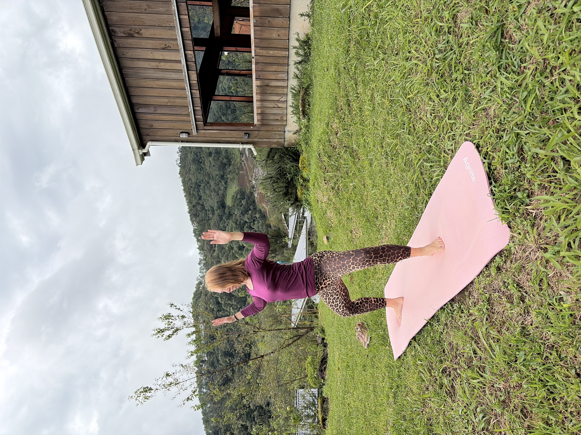 A medium-shot, full-body, eye-level, outdoor photograph shows a woman performing a yoga pose on a pink yoga mat placed on a grassy lawn. The woman has shoulder-length auburn hair and is wearing a long-sleeved, deep purple top and leopard-print leggings. Her arms are raised with palms facing forward, and her body is in a low warrior pose, with her right leg bent and forward. Her left leg is extended backward. In the background, a rustic, wooden building with large windows is visible to the right. Beyond the building and the woman, lush green hills and trees are seen under a cloudy sky, suggesting a rural or mountainous setting, possibly within the vicinity of Ban Khun Wang, Thailand, which is known for its natural beauty and agricultural landscapes. A pair of tan athletic shoes are placed on the grass near the yoga mat to the left.