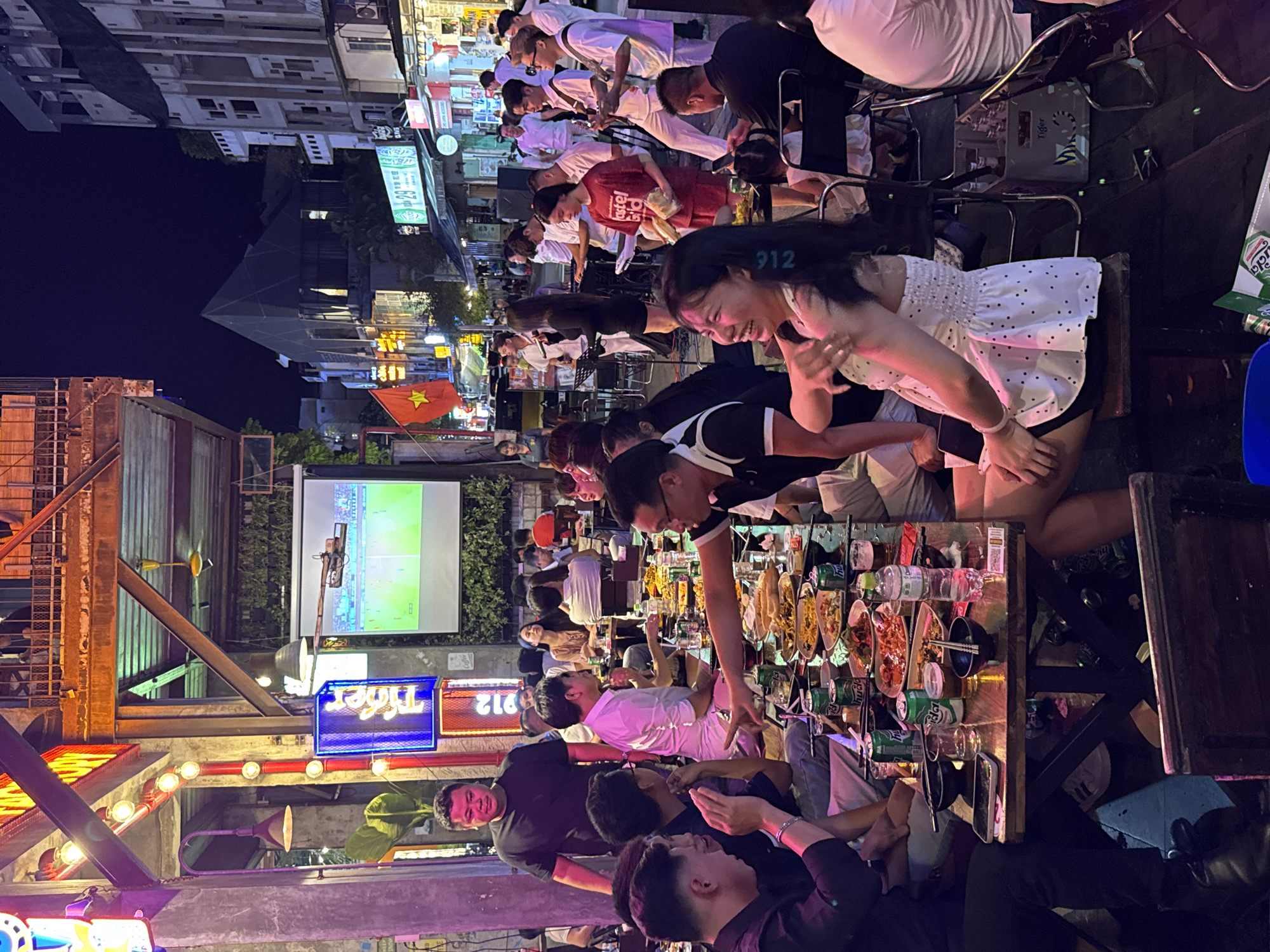 The image depicts a lively outdoor dining scene at night in Thuận Hóa Ward, Vietnam, with a group of people gathered around tables laden with food and drinks. A band is performing on a stage in the background, with several musicians playing instruments. A large projection screen displays a sports event, adding to the festive atmosphere. The area is illuminated by neon signs and string lights, creating a vibrant and energetic ambiance. The people are engaged in conversation and enjoying their meals, suggesting a social gathering or celebration. The scene captures the essence of a night out in Vietnam, with its blend of food, music, and entertainment.