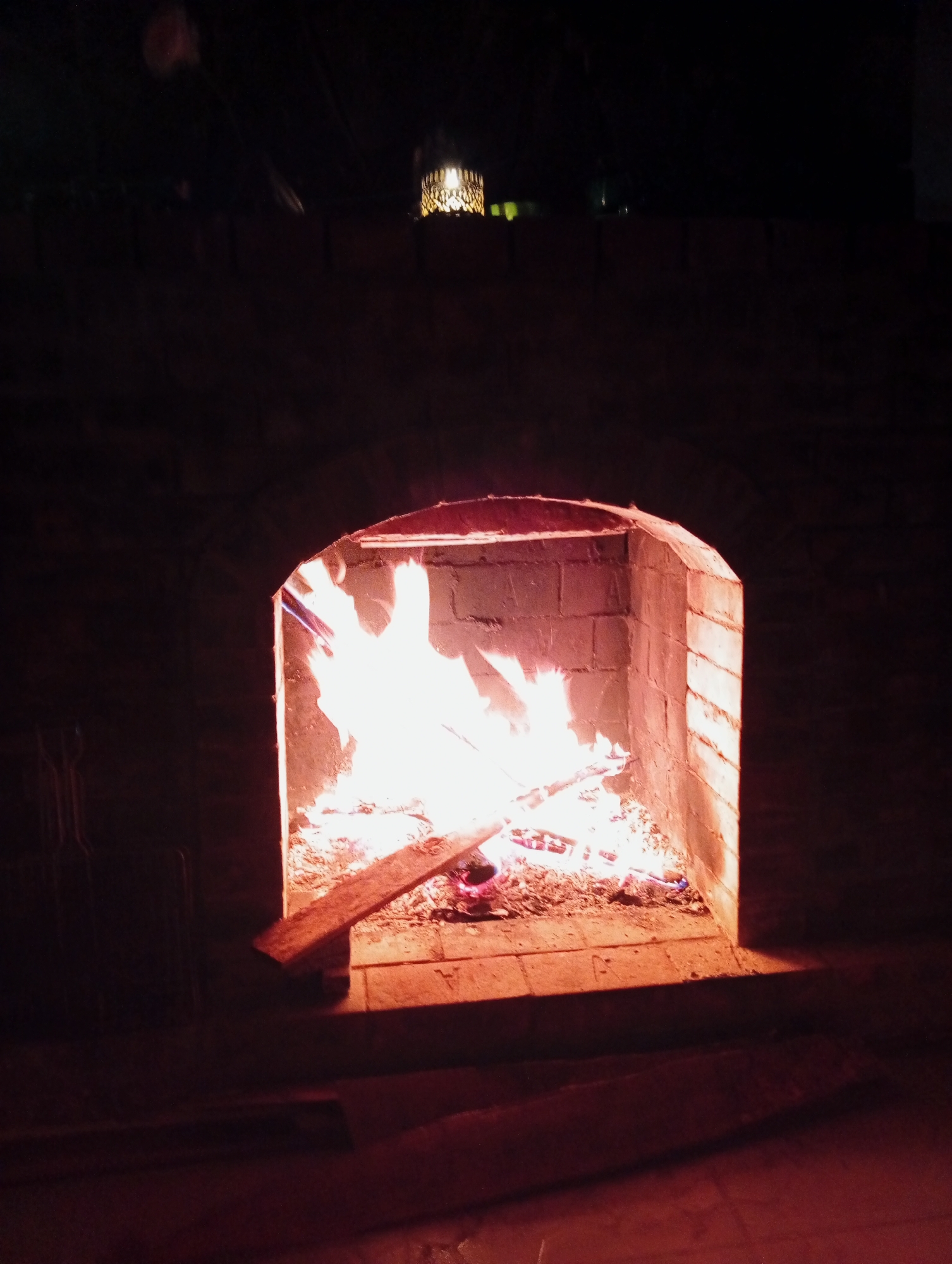 The image shows a brick fireplace with a roaring fire inside. The fire is consuming wood and producing bright flames. The fireplace is built of brick and has a simple arched opening. Outside the fire is a dimly lit area, possibly a room or outdoor space. The location in Lạc Dương, Vietnam is unspecified and not visually verifiable from the image. No people are visible in the image.